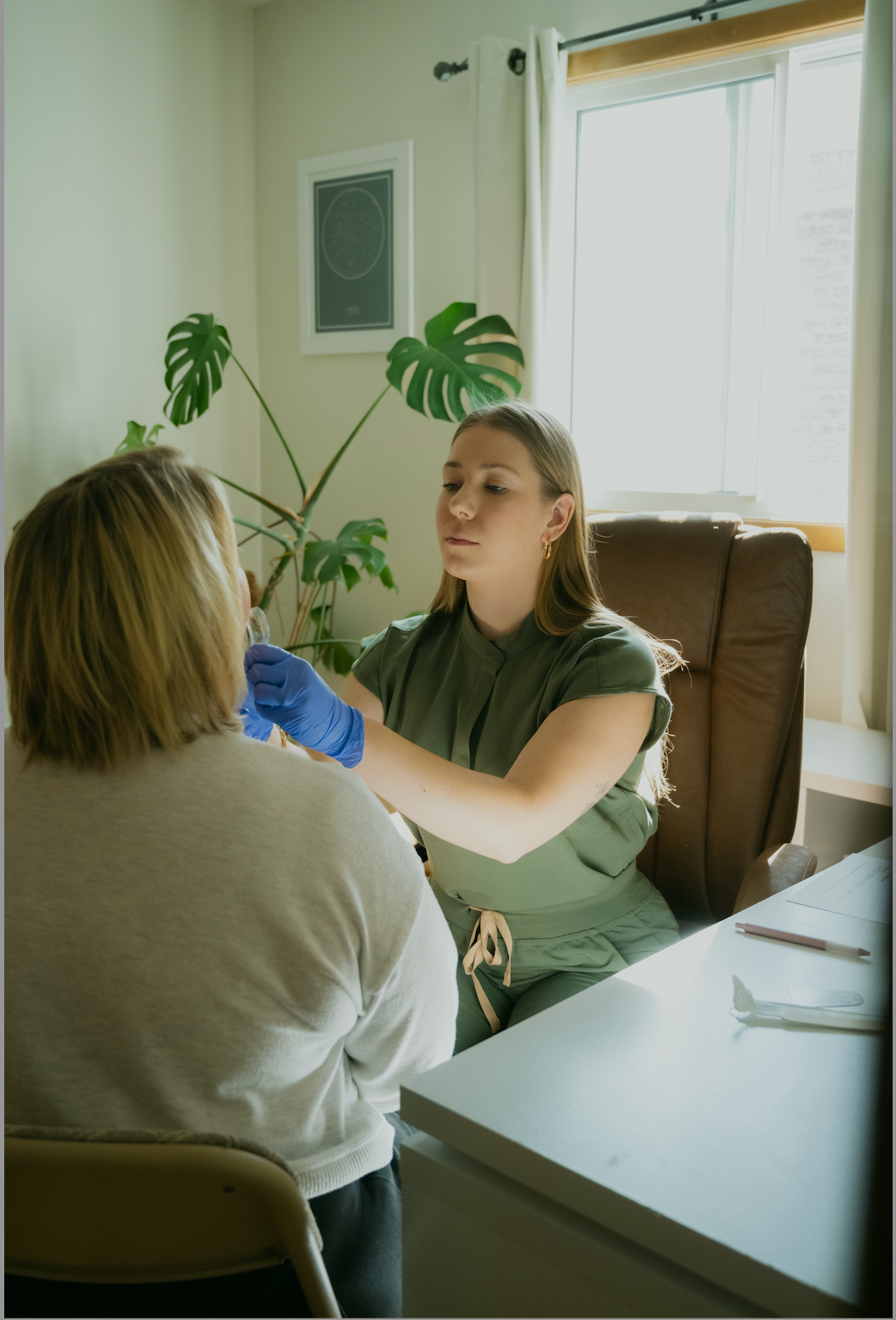 A healthcare professional in green scrubs and blue gloves administering a test to a woman with blonde hair in an indoor setting with a houseplant and a window in the background.