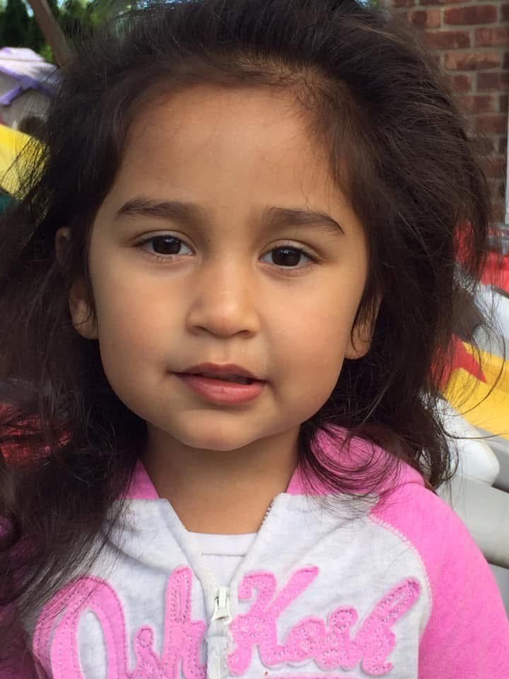 A young girl with long, dark, wavy hair and tan skin wearing a pink and white hoodie.