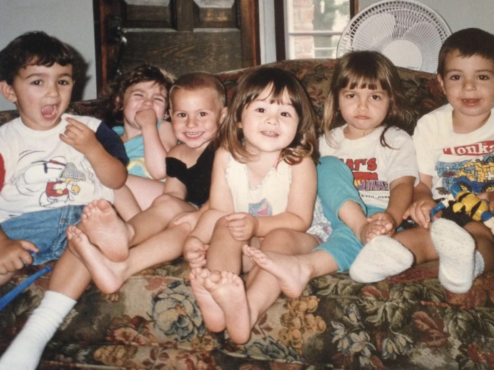 Seven young children sitting and lying on a floral couch, smiling and making playful expressions. They are in a room with a window and a fan in the background.