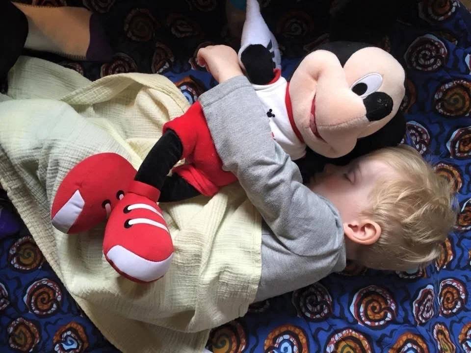 A young child lying in bed hugging a large Mickey Mouse plush toy.
