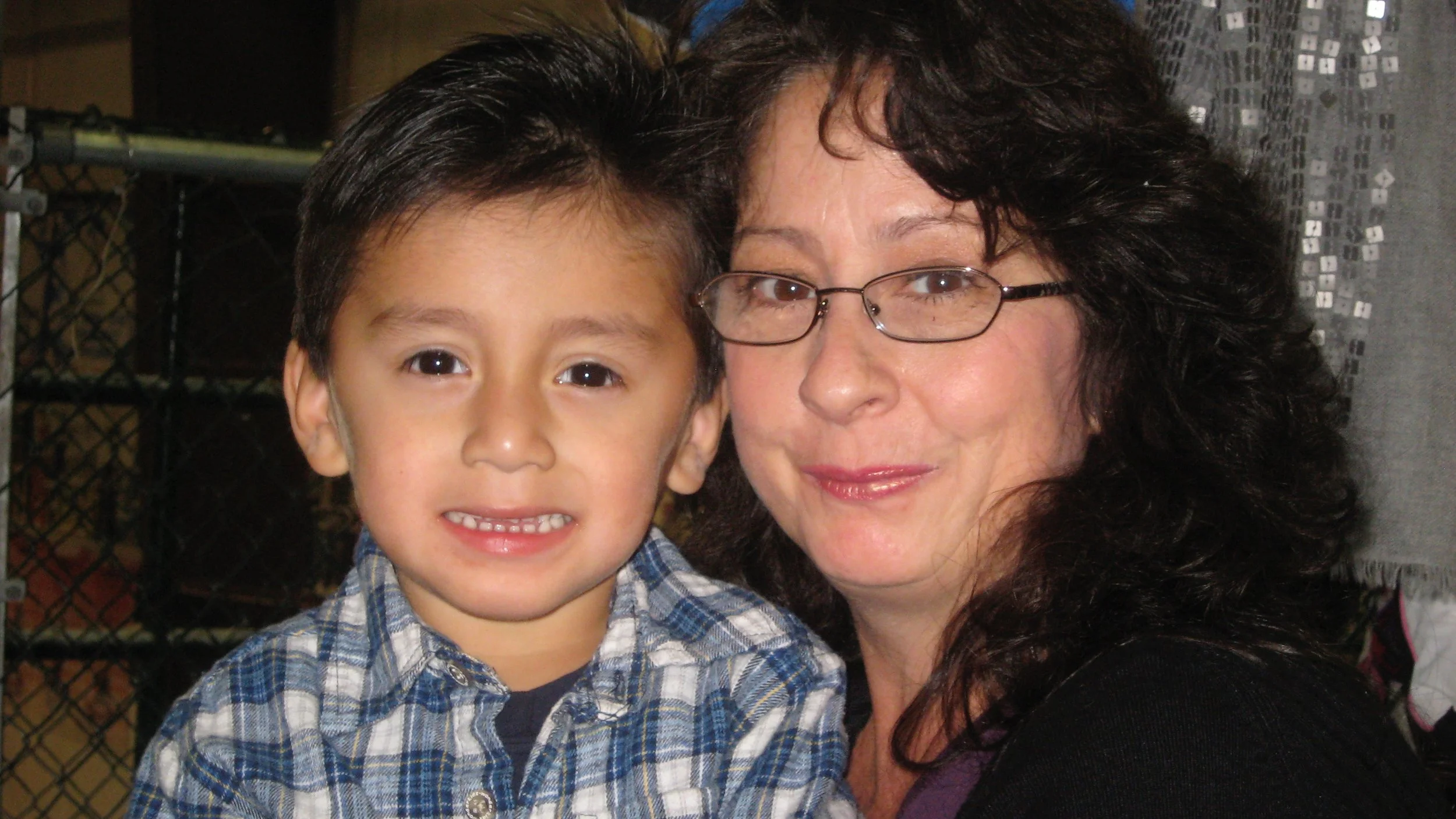 A woman with glasses and curly dark hair smiling beside a young boy with dark hair, brown eyes, and a plaid shirt, both looking at the camera.
