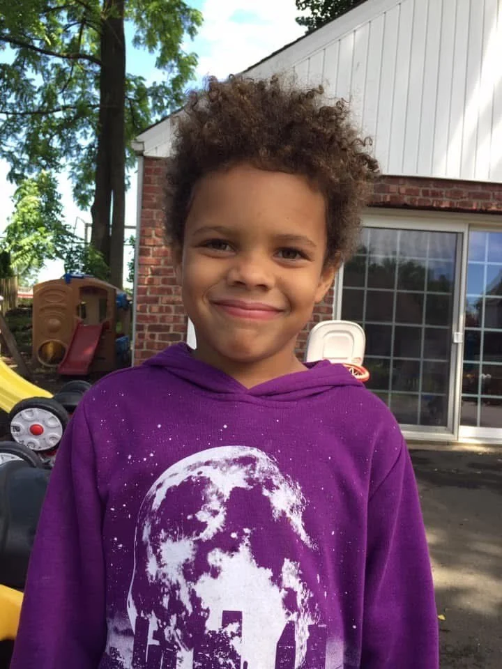 A young boy with curly hair, smiling, wearing a purple hoodie with a graphic of the moon printed on it, standing outdoors near a brick house with a sliding glass door, trees, and playground equipment in the background.