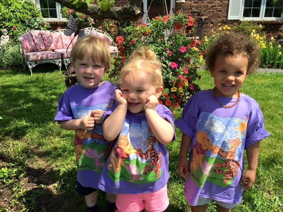 Three young children standing outdoors in a garden with flowers, wearing matching purple shirts with cartoon characters, smiling and posing for the photo.