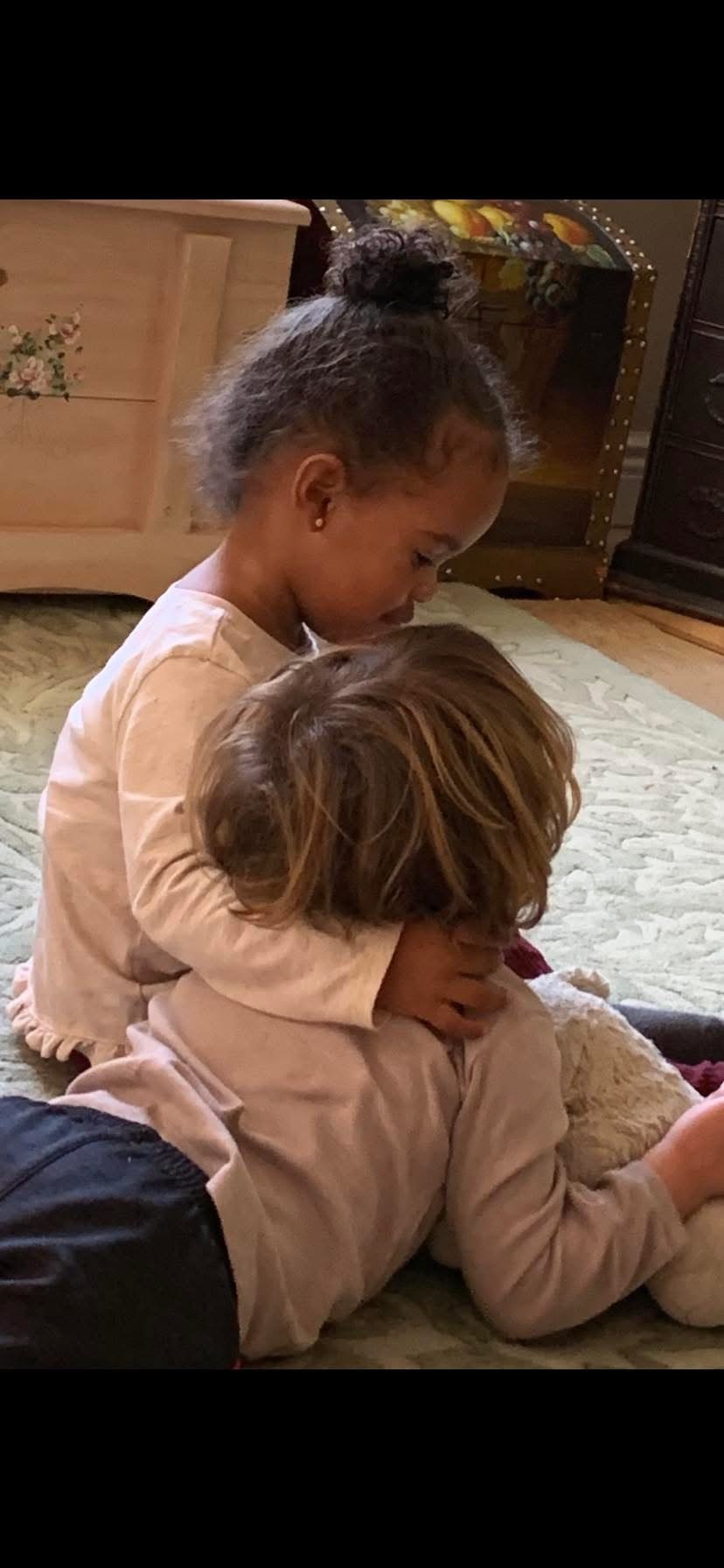 Two young children, a girl with a top bun hairstyle and a boy with long wavy hair, sitting on a quilted bedspread indoors, with the girl hugging the boy from behind.