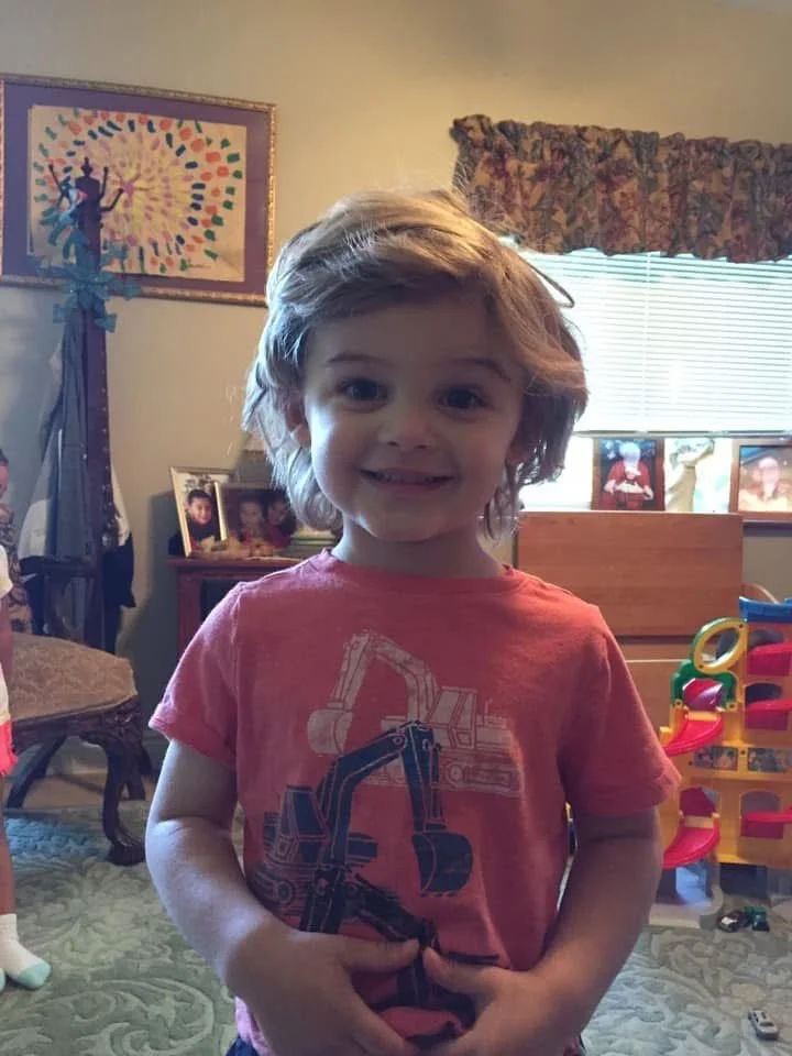Young girl with wavy hair smiling at the camera, wearing a pink t-shirt with construction equipment graphic, standing in a living room with framed photos, a window with a floral valance, and colorful toy storage in the background.