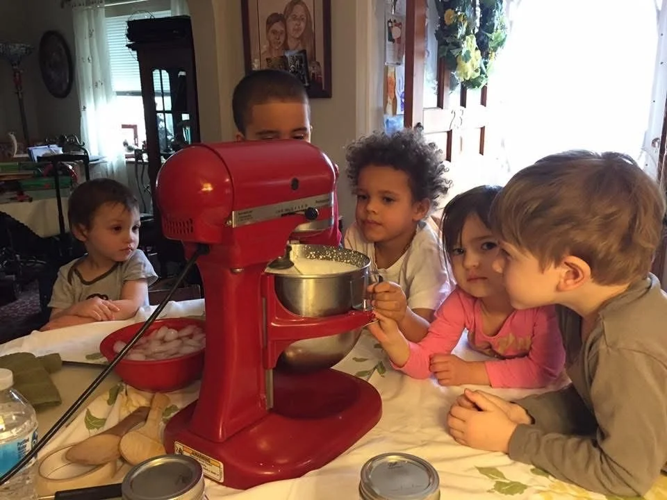 Children gathered around a red stand mixer, with one child operating it, while others watch and wait. The scene takes place at a kitchen table, with jars, a bowl of ice, and other kitchen items visible.