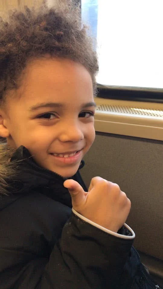 A young boy with curly hair smiling and giving a thumbs-up, sitting next to a window on a bus or train.