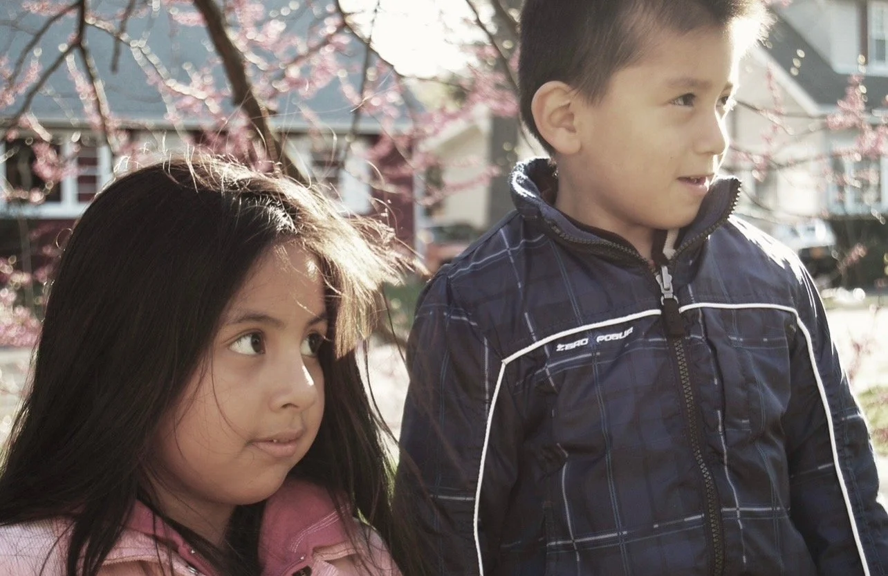 Two young children, a girl and a boy, standing outdoors near blooming pink cherry blossom trees. The girl has long dark hair, and the boy has short dark hair and is wearing a dark blue jacket. They are looking in different directions with sunlight illuminating the scene.