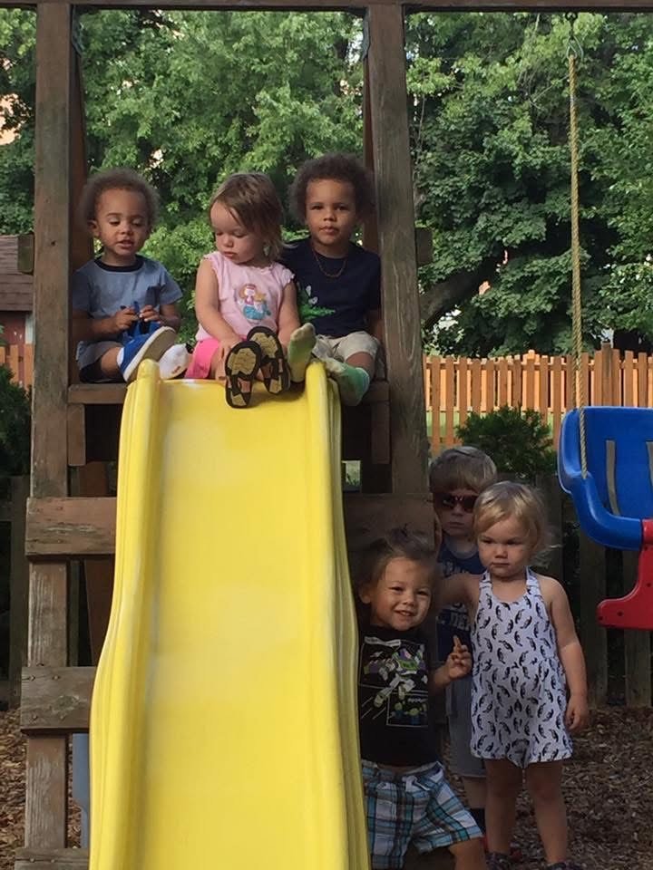 Six young children on a playground structure with a yellow slide, with trees and a wooden fence in the background.