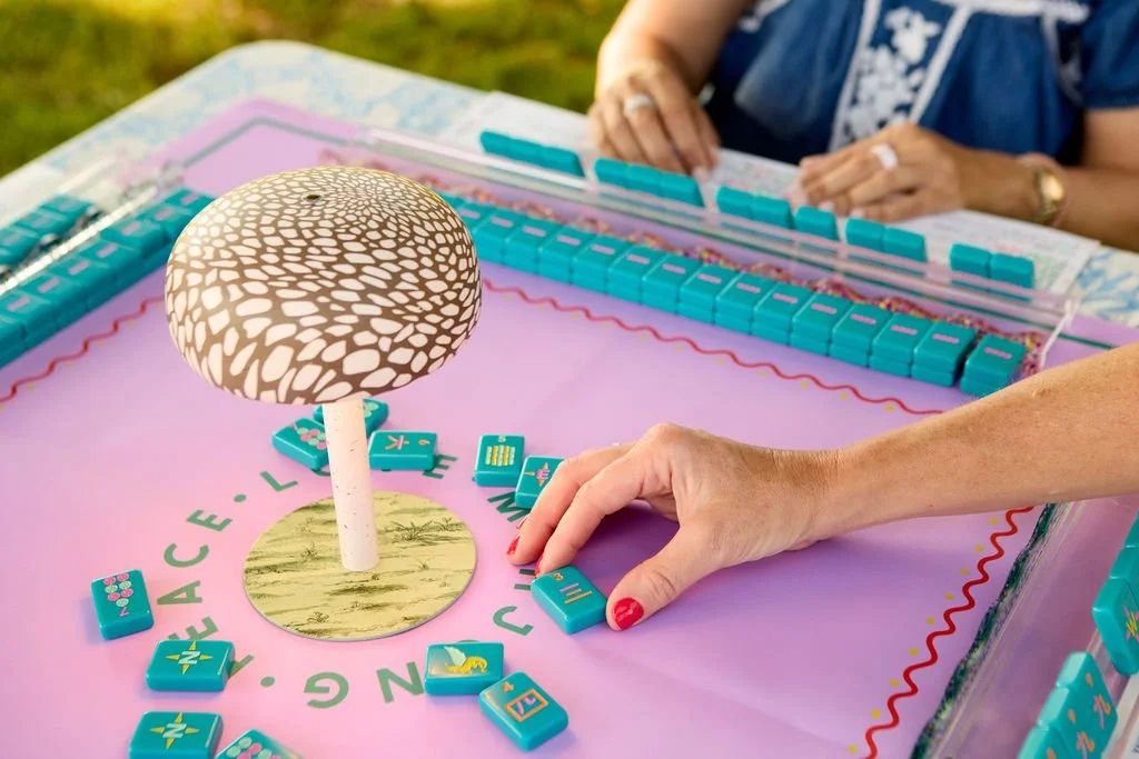 Close-up of a domino game in progress on a pink table, with a decorative mushroom centerpiece. Two women are seen playing, one reaching for a domino, outdoors on a sunny day.