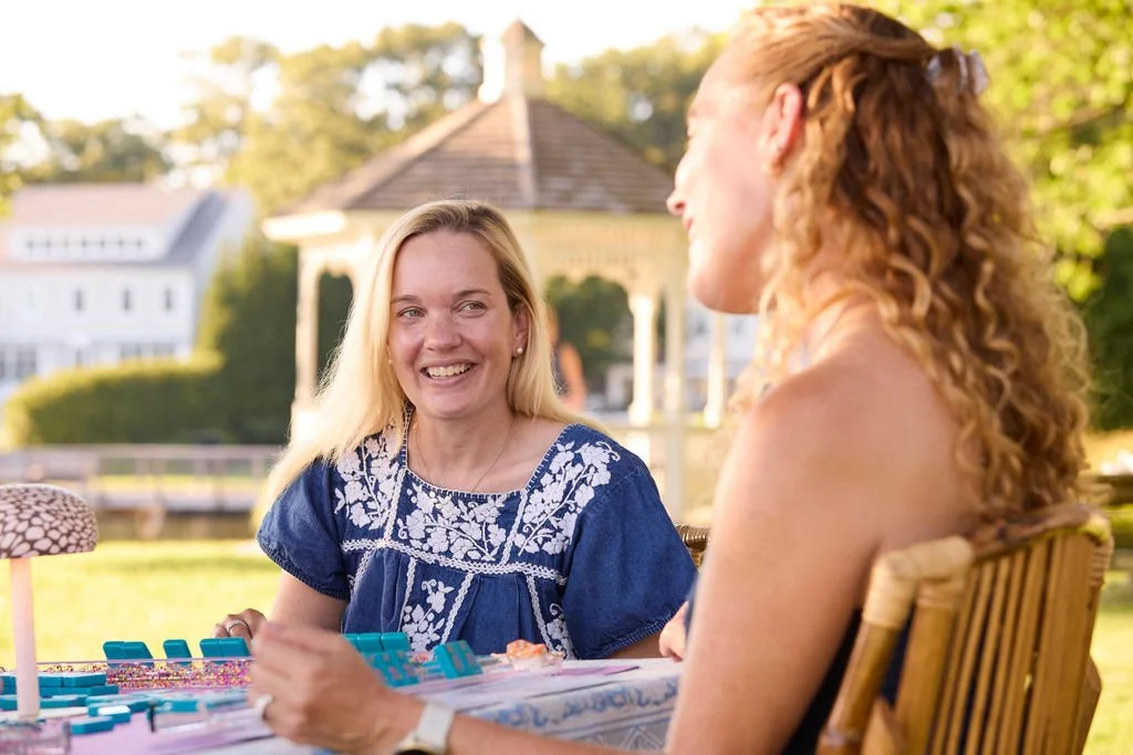 Two women sitting outdoors at a table, smiling and talking, with a gazebo and trees in the background, in a sunny setting.