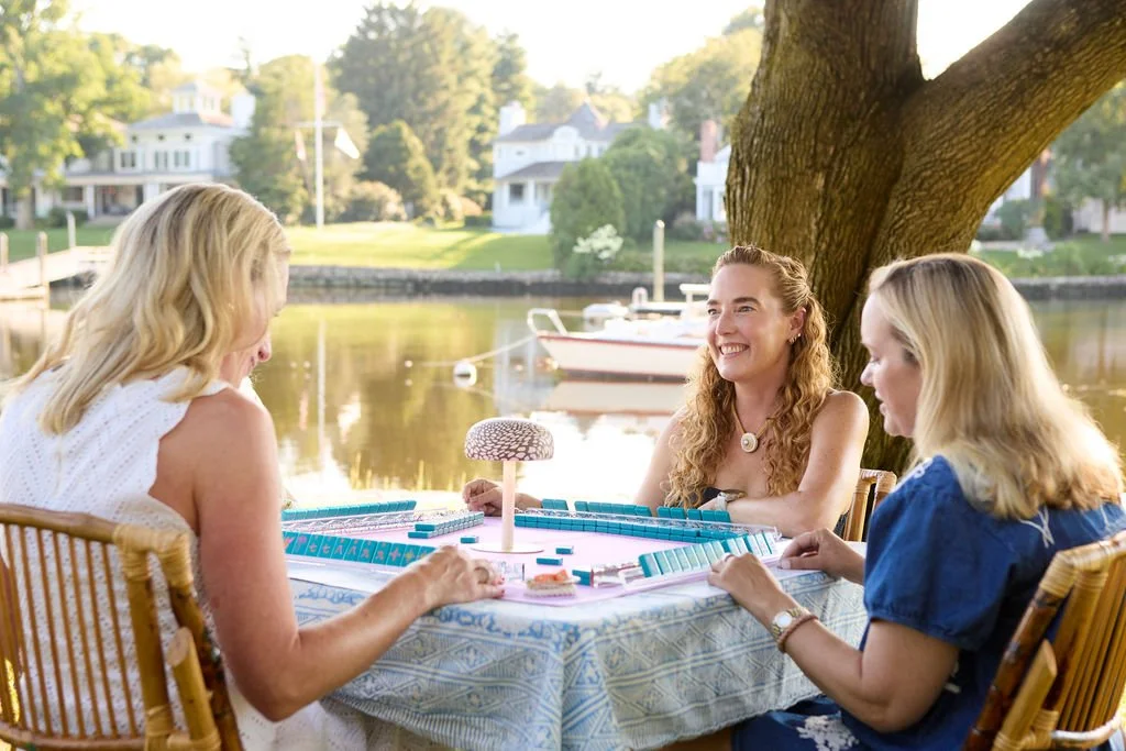 Three women playing Monopoly by a river with boats, trees, and houses in the background on a sunny day.