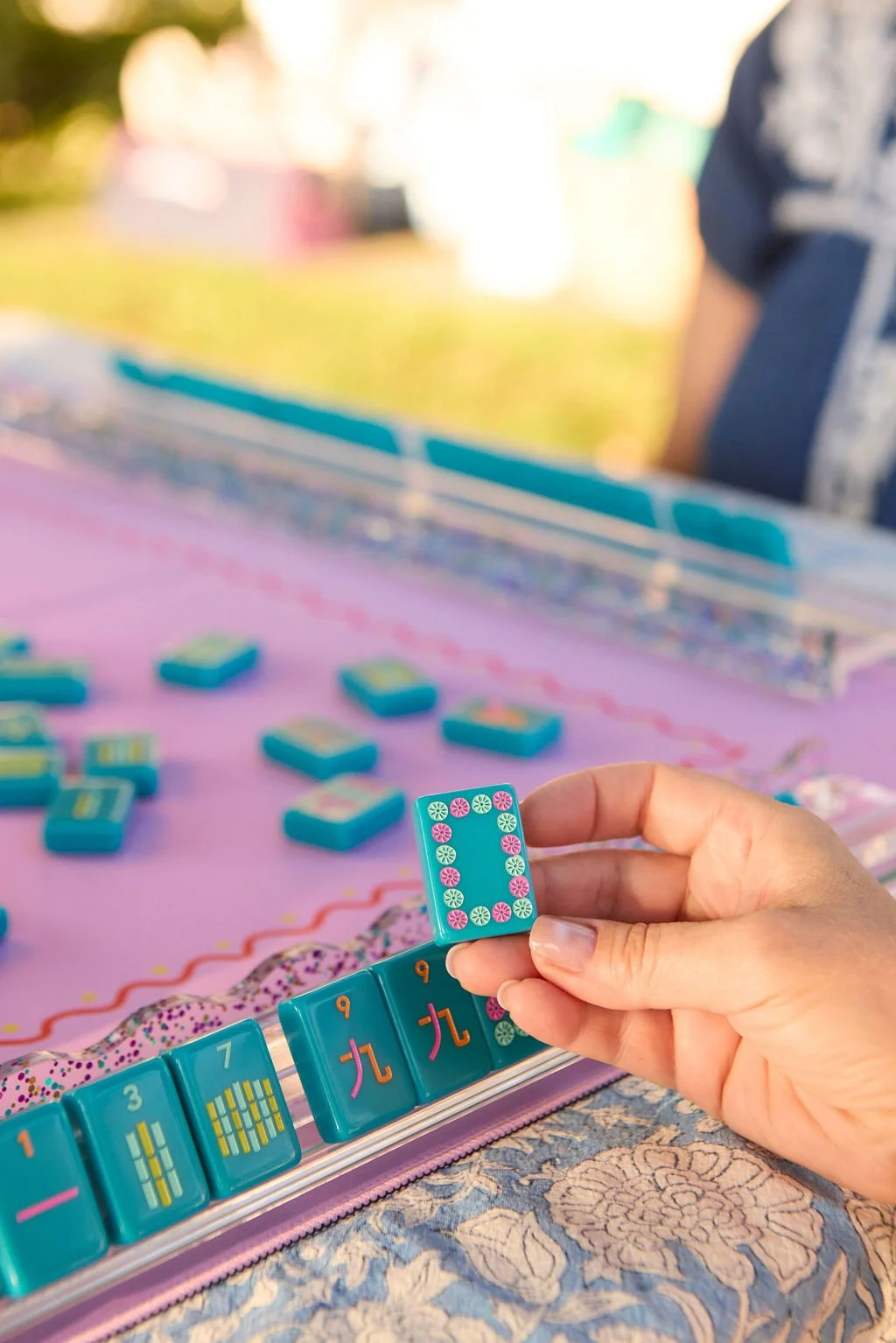 Person holding a Mahjong tile with a pink and green flower design at a Mahjong table with blue tiles, some showing numbers and patterns, on a pink playing surface.