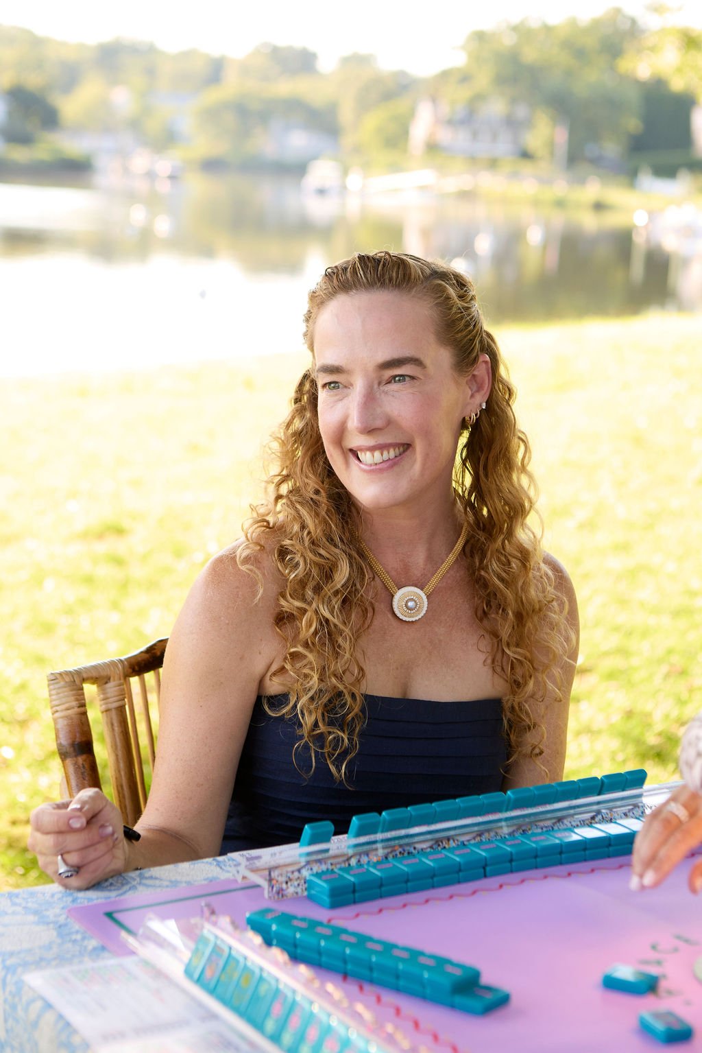 A woman with curly red hair, wearing a black strapless top and a necklace, playing a game of dominoes outdoors by a lake, smiling.