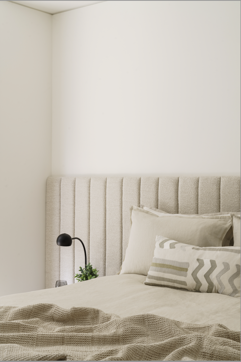 A neatly made bed with beige linens, decorative pillows, an upholstered beige headboard, a black bedside lamp, and a small green plant on the nightstand. The room has white walls and a calm, neutral decor.