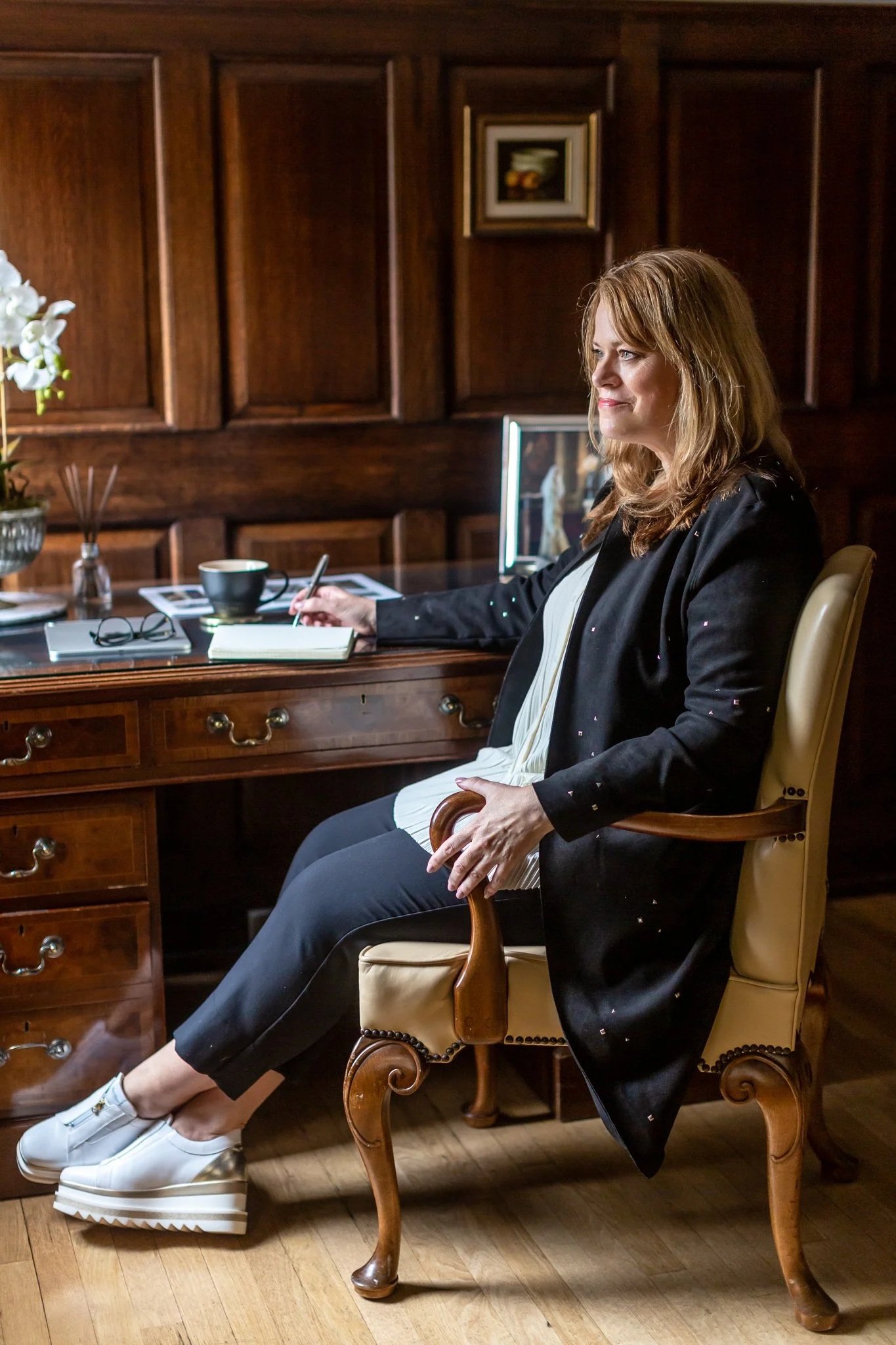 A woman sitting on an ornate wooden chair in a room with dark wooden paneling, writing in a notebook at a desk with various items, including a coffee cup, glasses, and a flower vase.