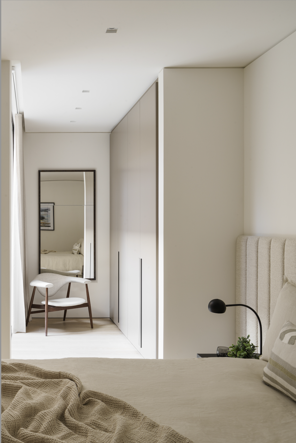 Minimalist bedroom with beige bedding, a black bedside lamp, a small potted plant, and a large mirror by the window. A chair with a curved back and wooden legs is near a wall with closet doors.