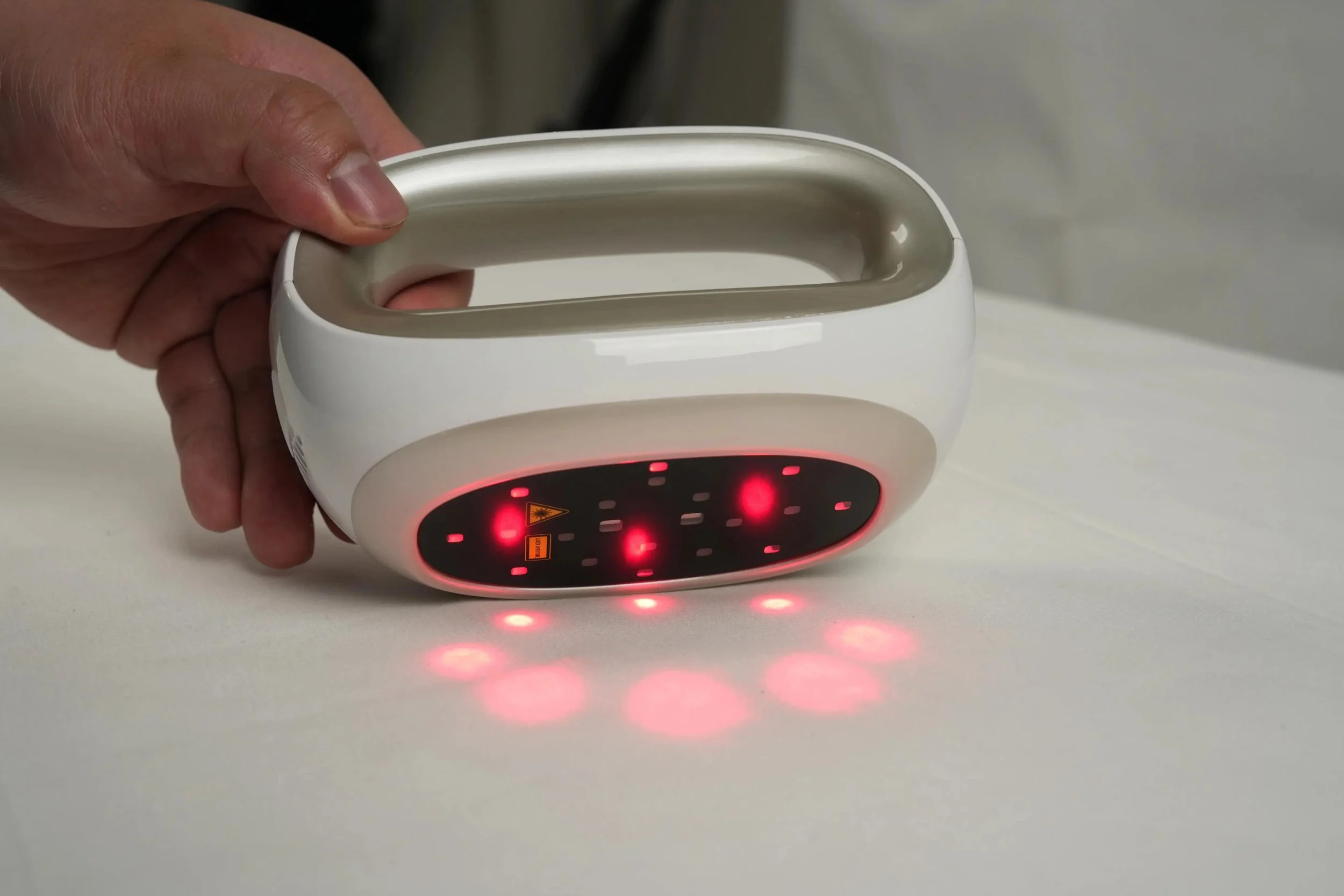 A person holding a white device with a black display panel that has red lights and symbols, emitting red laser-like dots onto a white surface.