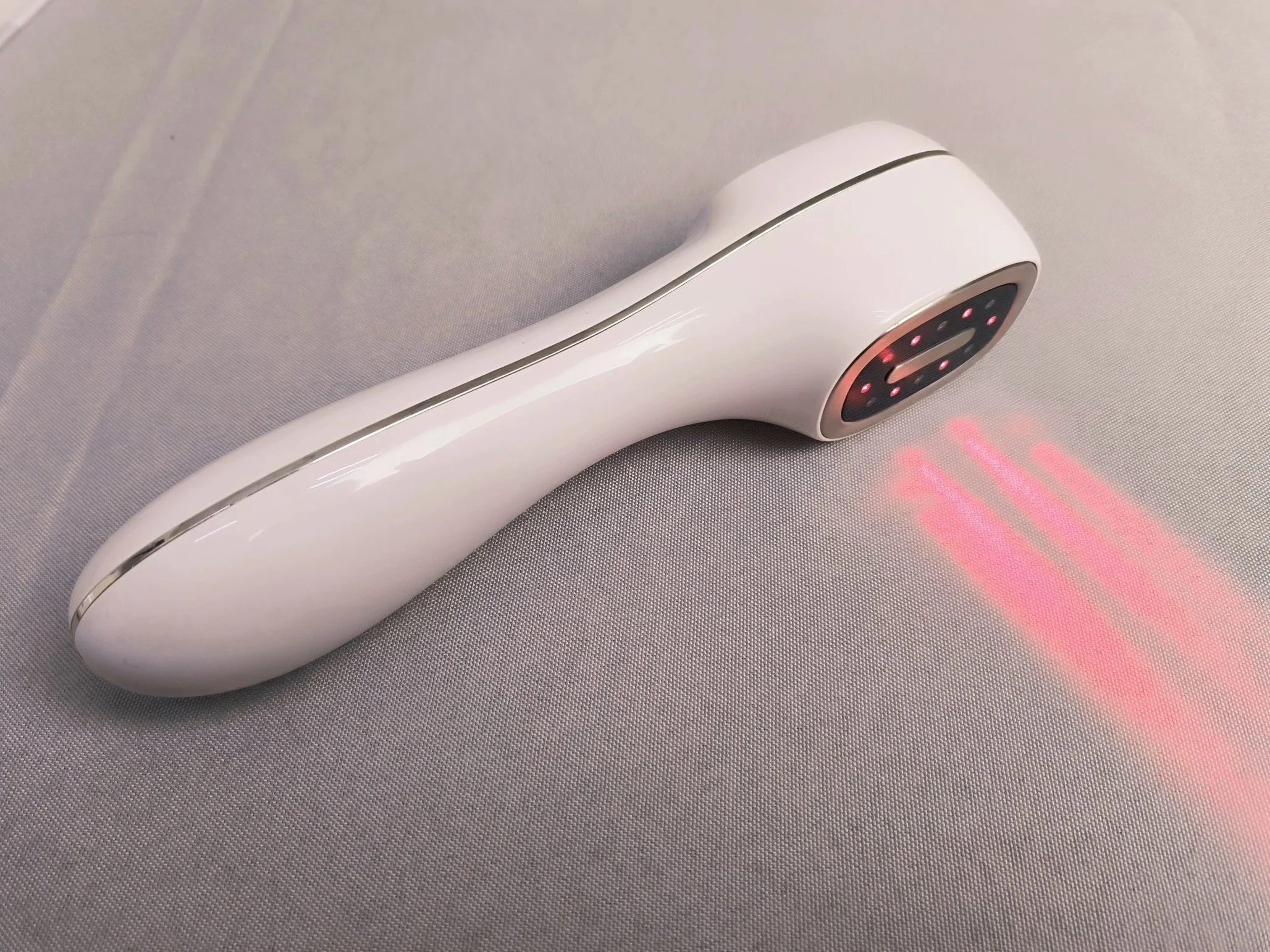 A handheld device, possibly a laser hair removal or skin treatment tool, with a white body and a black and red digital display, resting on a grey textured surface, with a red laser beam emanating from the tip.