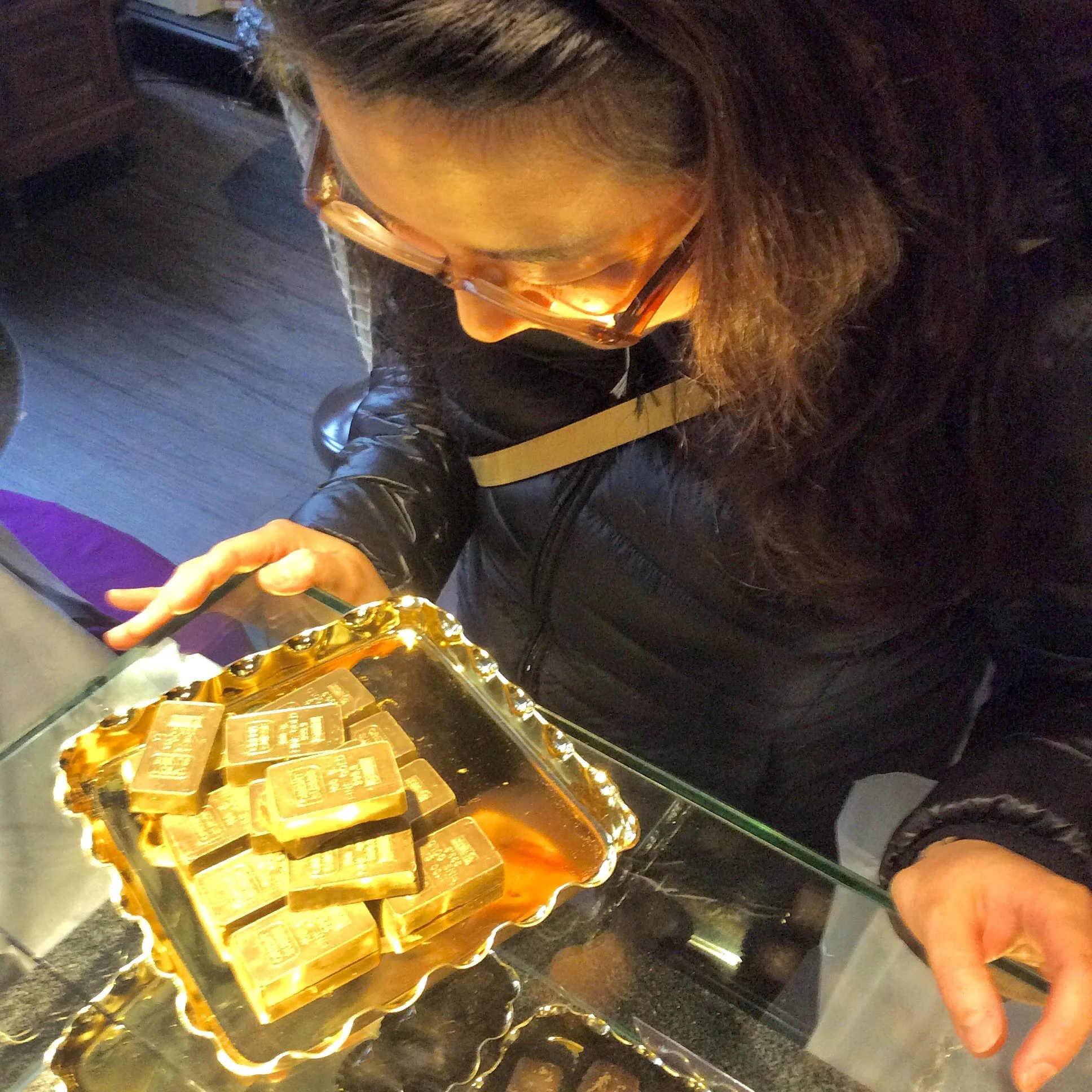 A person with glasses and long hair is holding a fork and serving a rectangular-shaped dish with gold edges on a platter, with several individual pieces arranged in a row. The setting appears to be in a restaurant or formal dining environment.