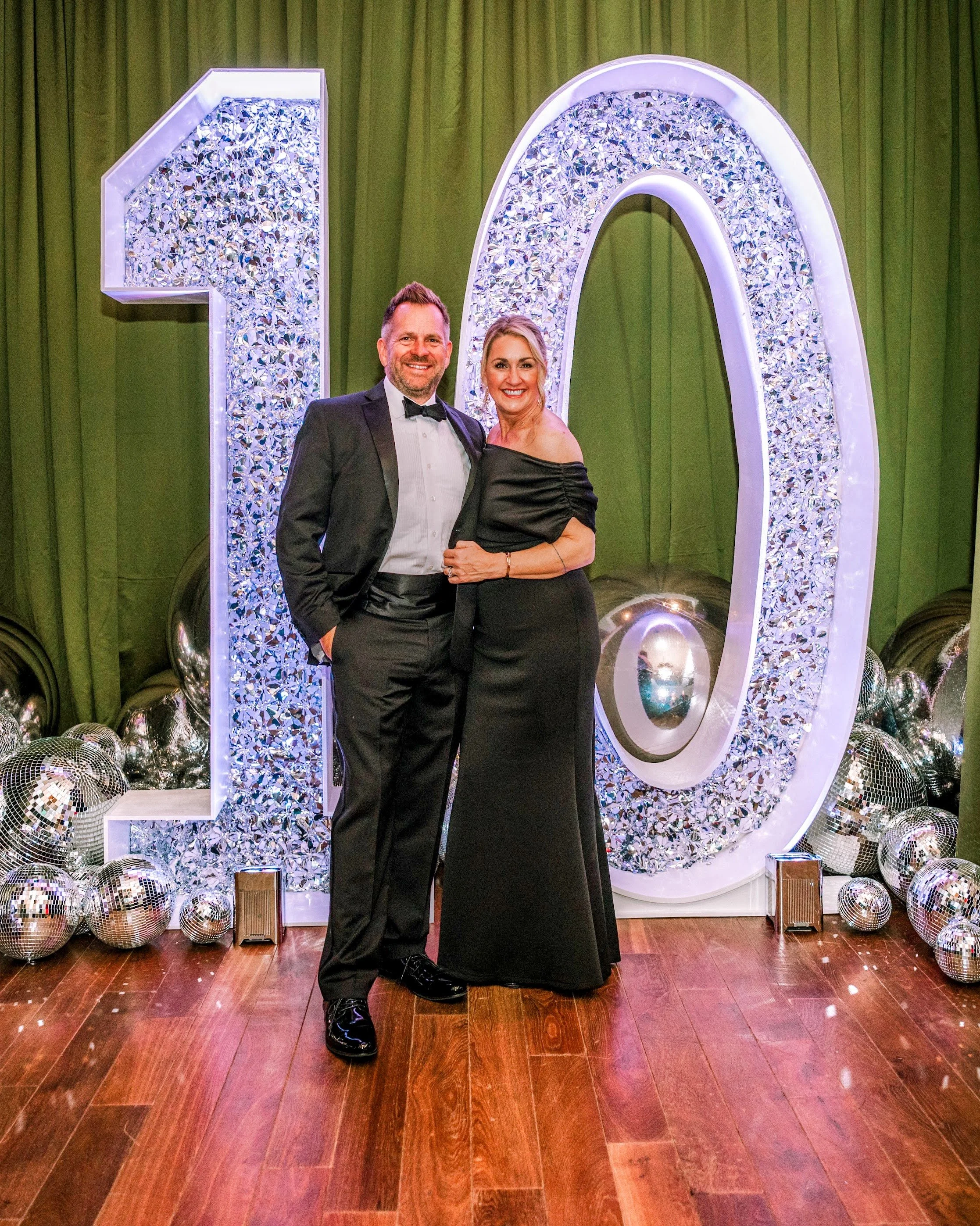 A man and woman dressed in formal attire stand in front of a large illuminated number 10 with decorative details, green curtains, and scattered disco balls.