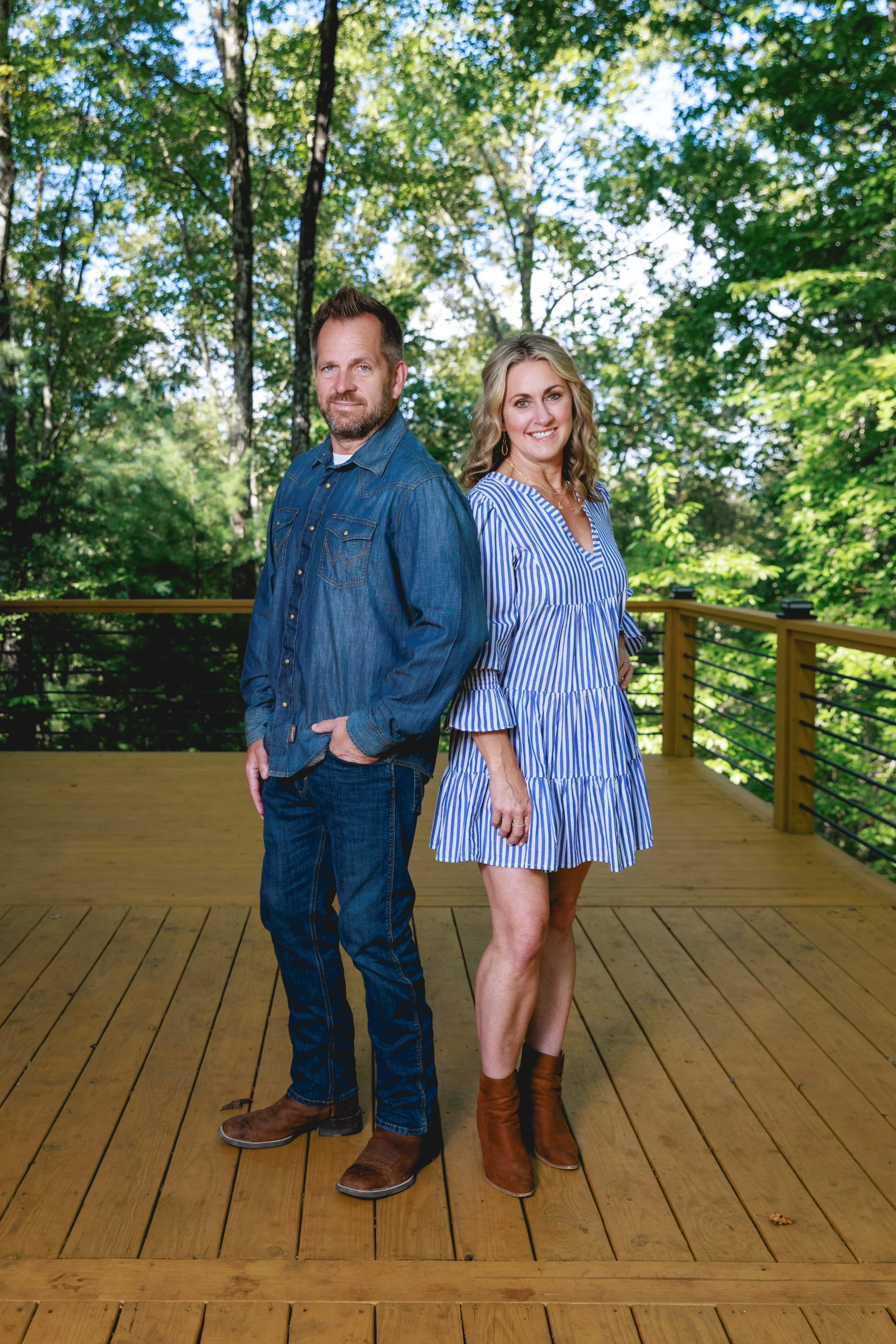 A man and woman stand back-to-back on a wooden deck with green trees in the background. The man wears a denim jacket and jeans, and the woman wears a blue and white striped dress with ankle boots. Both are smiling softly.