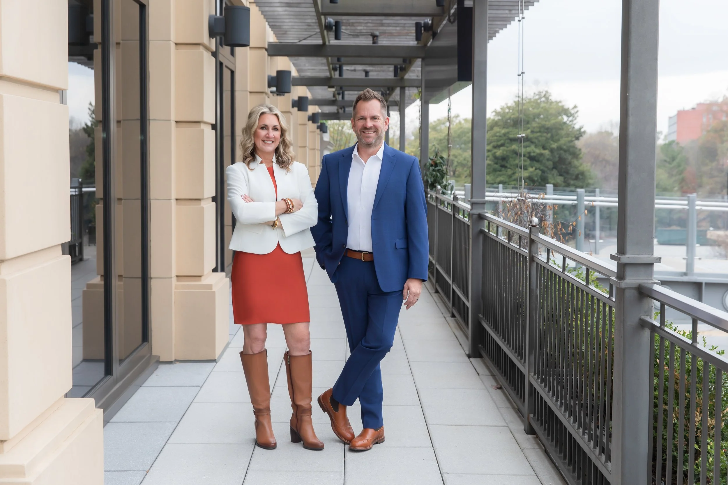 A woman and a man standing on a balcony, smiling at the camera. The woman is wearing a white blazer, orange dress, and brown boots. The man is wearing a blue suit, white shirt, and brown shoes. The background shows a cityscape with trees and buildings.