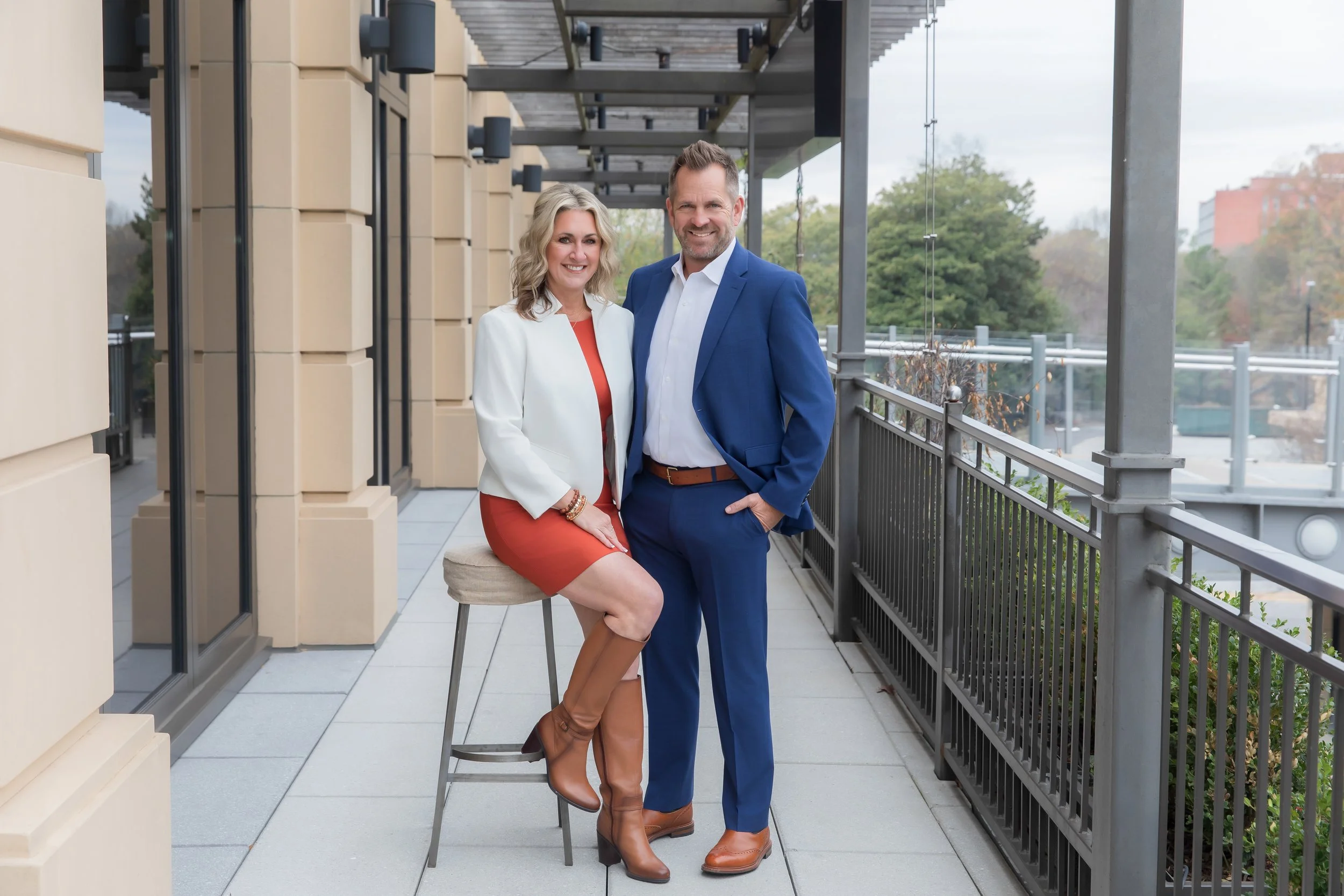 A woman in a white blazer and red dress sitting on a bar stool with knee-high brown boots, and a man in a blue suit standing next to her with one hand in his pocket, on a balcony with a city view and trees in the background.
