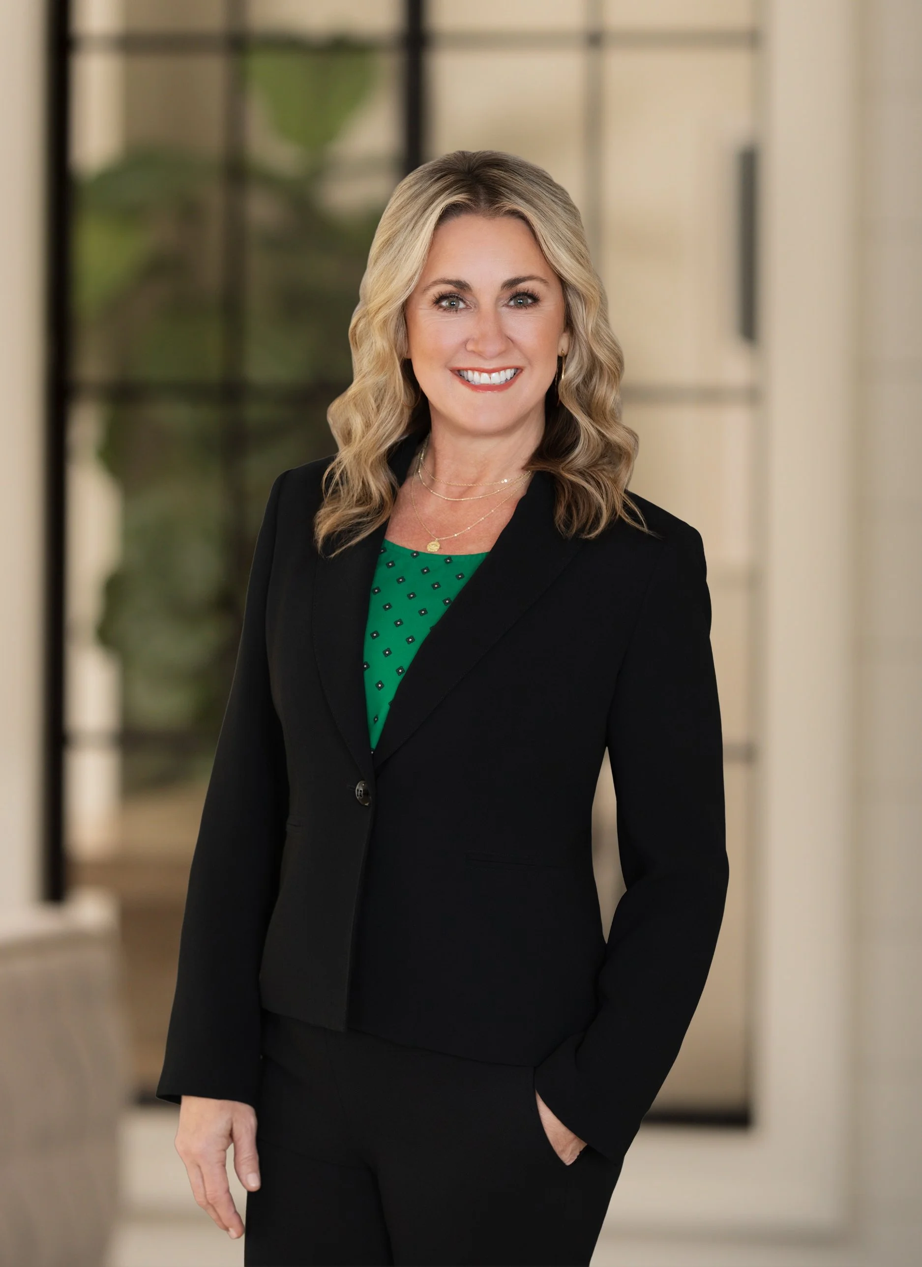 A woman with blonde wavy hair wearing a black blazer, green top, and gold jewelry, standing in a modern indoor space with a blurred background.