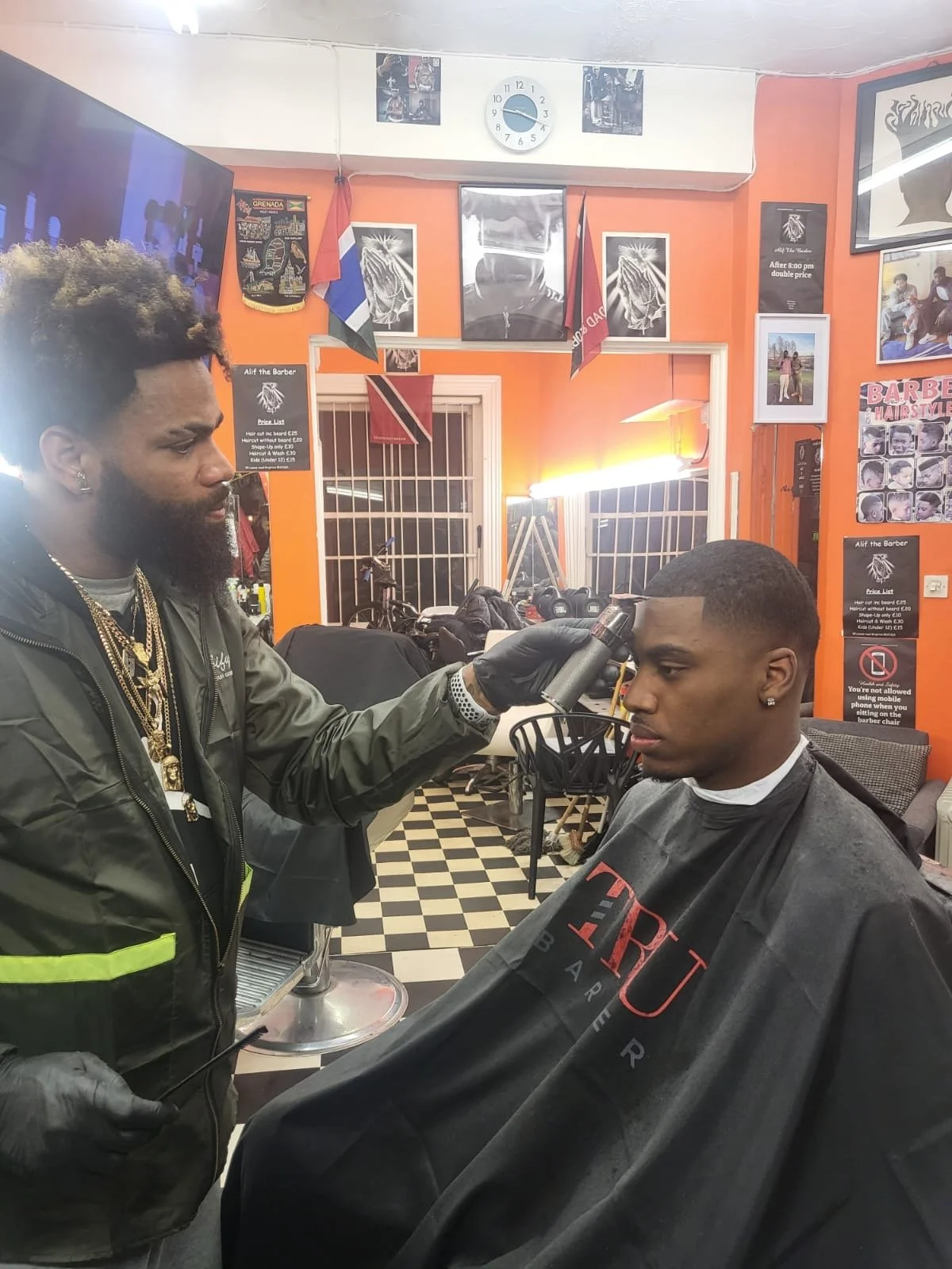 A barber giving a haircut to a man in a barbershop with orange walls, black and white checkered floor, and various posters and flags on the walls.