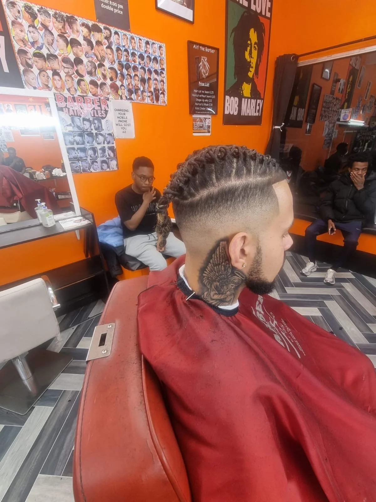 A young man with a braided hairstyle and tattoos on his neck sits in a barber shop, wearing a red barber cape. In the background, a boy with glasses and a black shirt is seated, with two other men seated on a bench, all in a brightly colored orange-w