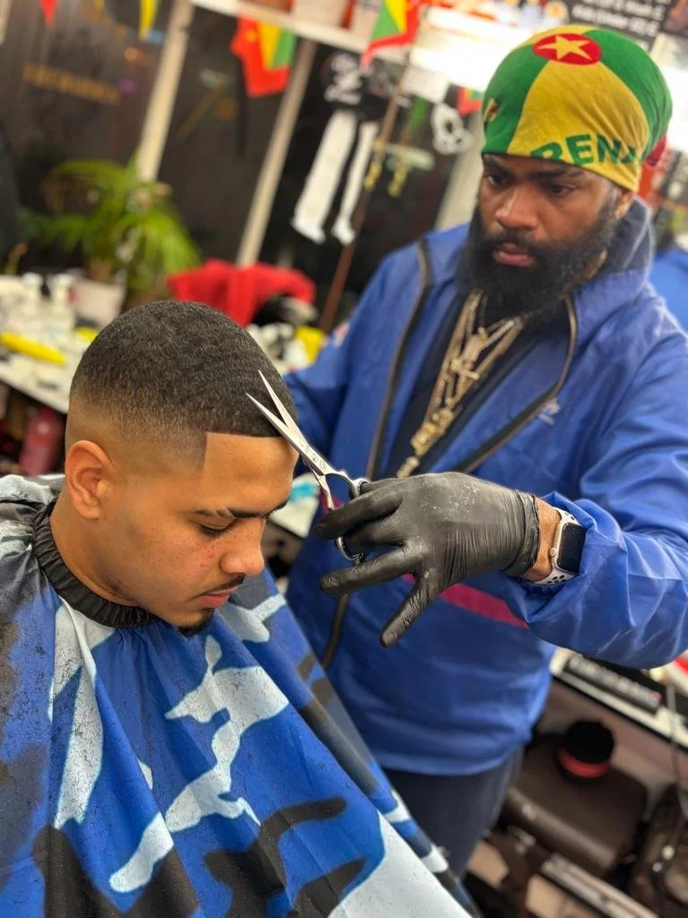 A man getting a haircut at a barbershop, with scissors in hand, while a barber with a bandana and black gloves is trimming the young man's hair.