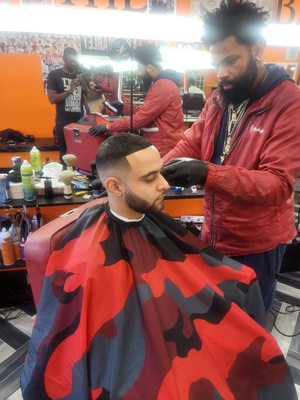 A man getting a fresh haircut at a barbershop, with a barber applying finishing touches. The customer is seated, wearing a red, black, and gray camouflage cape. The barbershop has orange walls, mirrors, hair products, and tools visible on the counter.