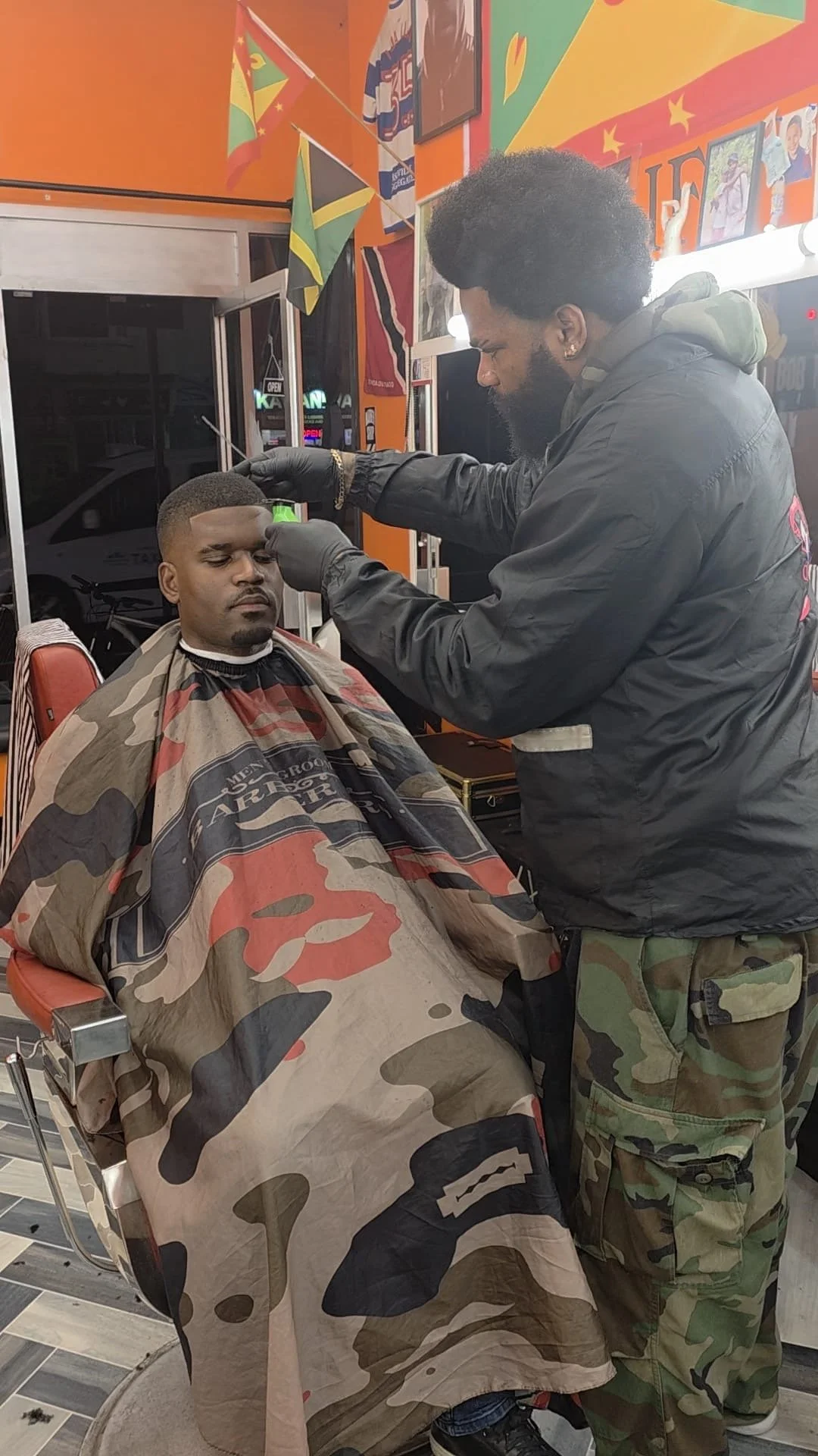 Man getting a fresh haircut at barbershop, seated with a camouflage cape, barber using clippers, colorful flags and posters on wall.