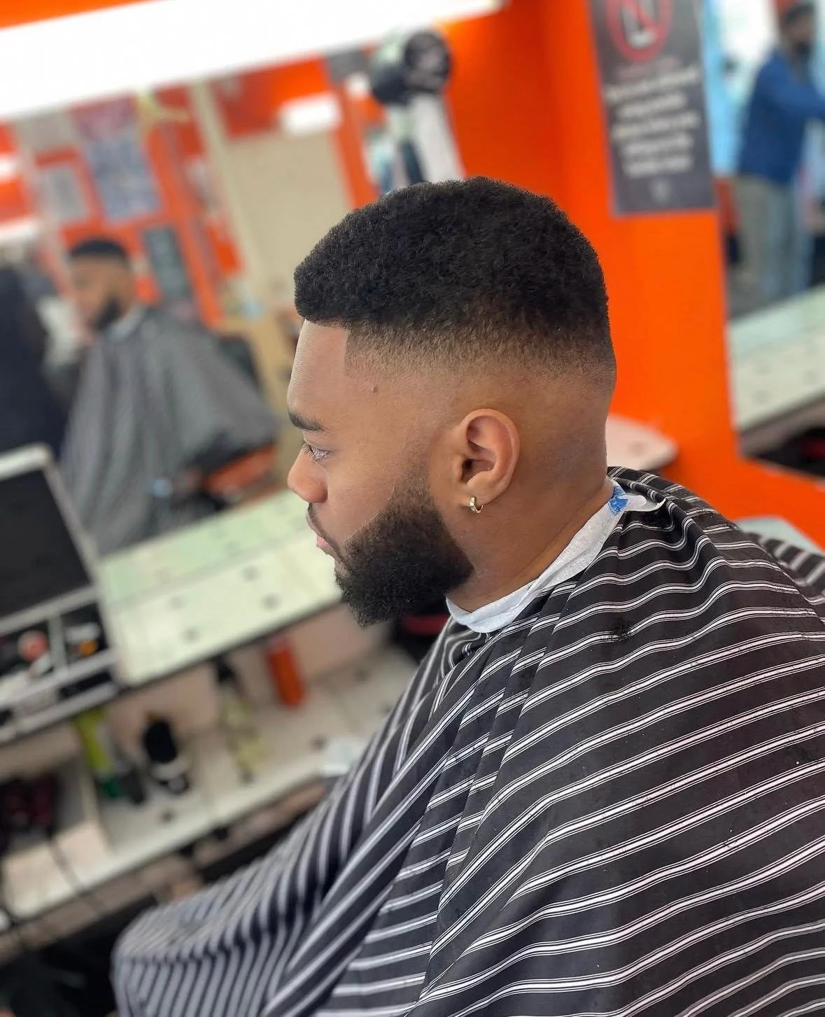 Young man with a fresh fade haircut, beard, and earring, sitting in barber shop with striped cape on, background shows other customers and barbers.