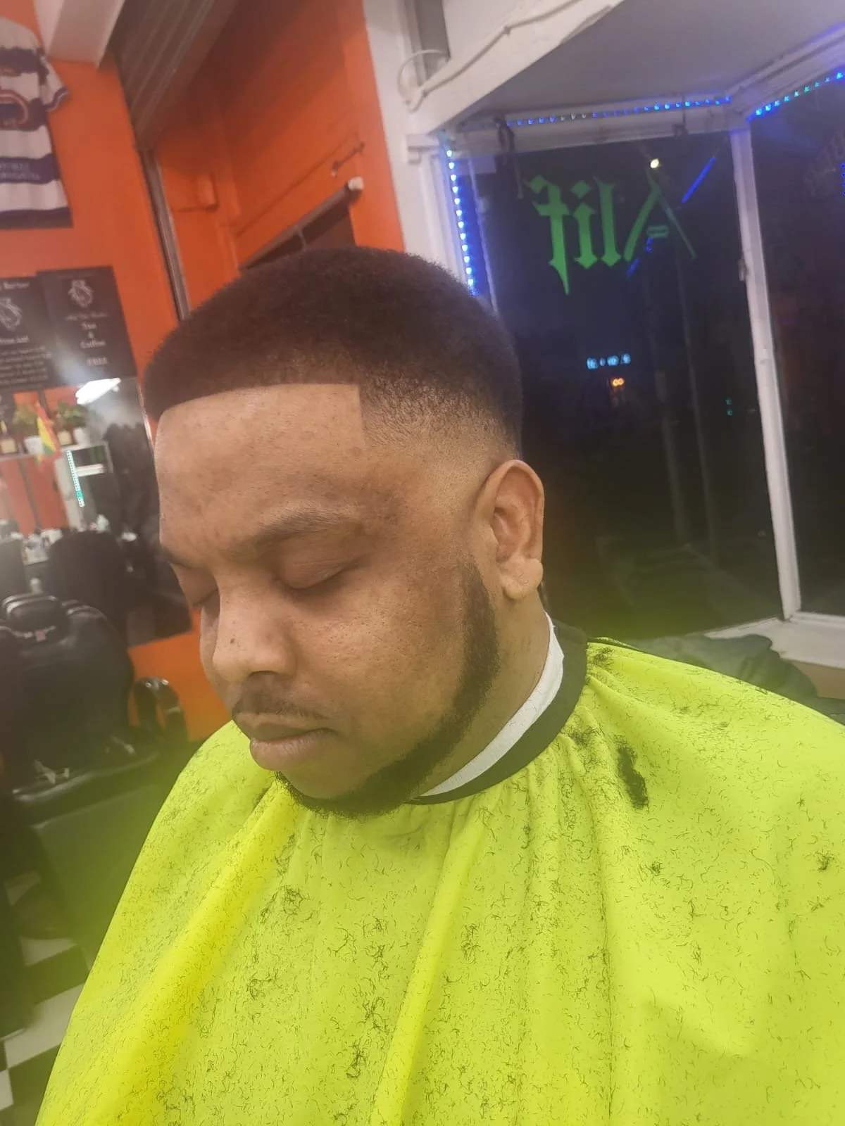 A man with a freshly cut fade hairstyle sitting in a barbershop, wearing a bright yellow cape, with closed eyes and a relaxed expression.