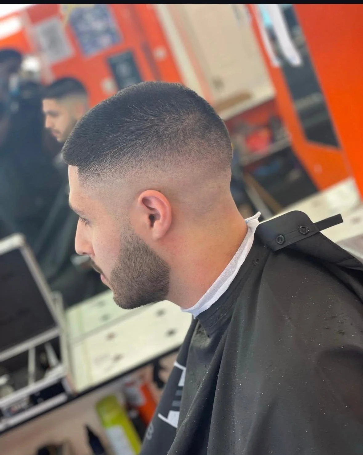 Man with a fresh skin fade haircut, beard, wearing a barber cape, in a barbershop with others and barber tools visible in the background.