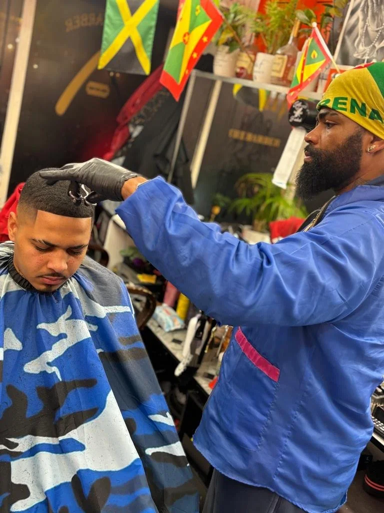 A barber giving a haircut to a customer in a shop decorated with Jamaican flags.