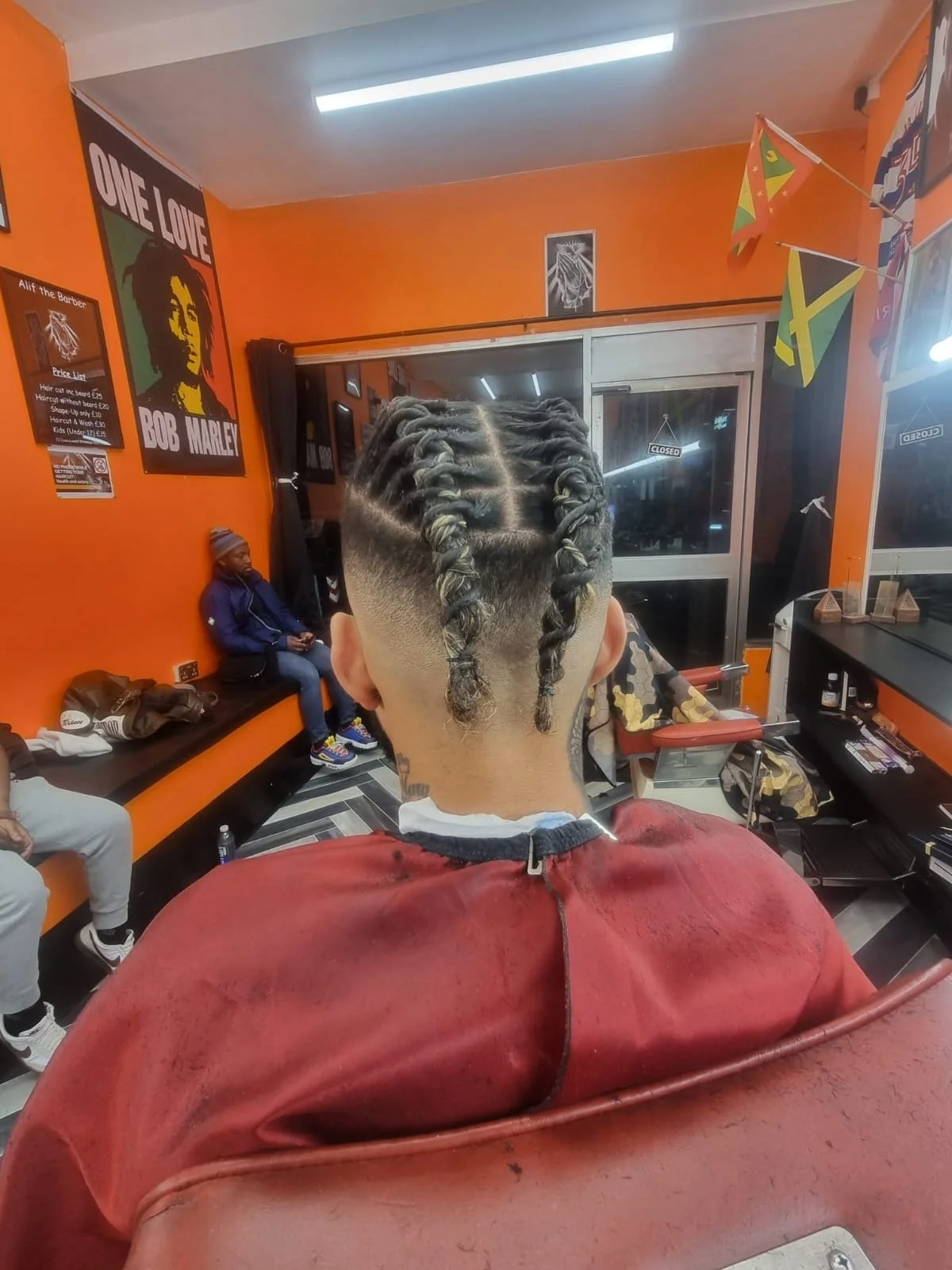 A person in a barbershop with a fresh hairstyle featuring cornrows and a fade. The barbershop has orange walls, various posters including a Bob Marley poster, and Jamaican flags. The person is seated with their back to the camera, wearing a red barbe