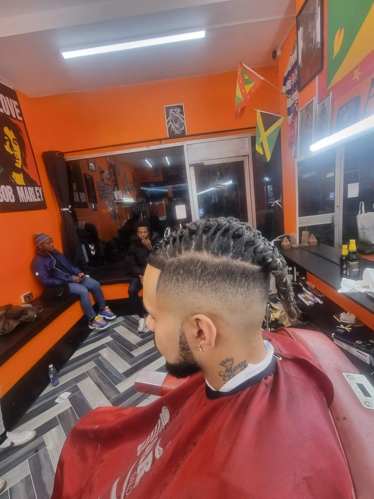 A man with a fresh braided hairstyle and a tattoo on his neck sits in a barber shop with orange walls decorated with Bob Marley posters and Jamaican flags. Two children are seated on a black bench, one wearing a hoodie and the other in a jacket. The 