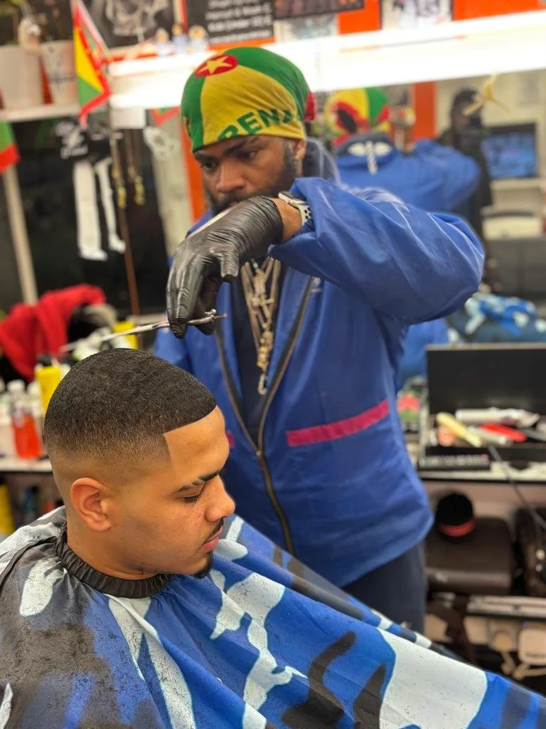 Barber giving a haircut to a young man in a barbershop, with the barber wearing gloves and a colorful headscarf.