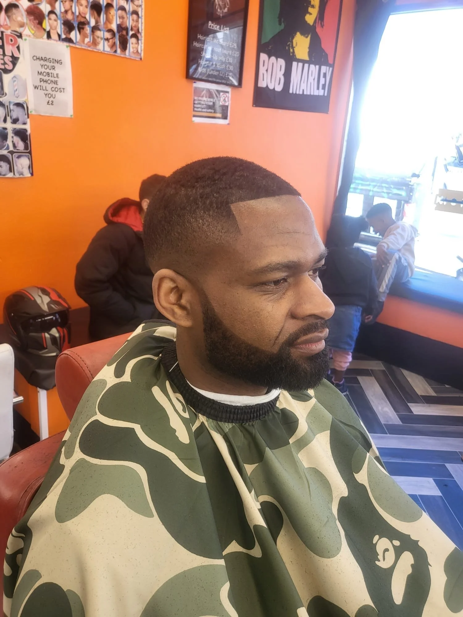 A man with a freshly cut hairstyle and beard seated in a barbershop, wearing a camouflage barber cape. There are children getting haircuts in the background, and the walls are decorated with posters, including Bob Marley.