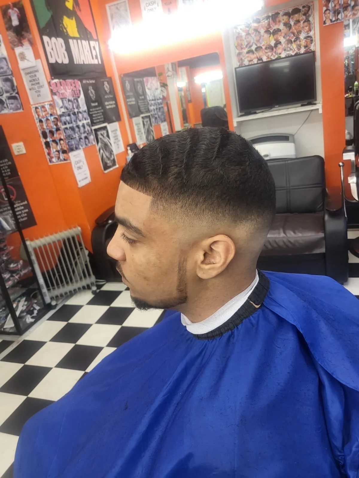 Young man with a fresh haircut sitting in a barber shop with orange walls and black-and-white checkered floor, surrounded by posters and pictures related to haircuts and hairstyles.