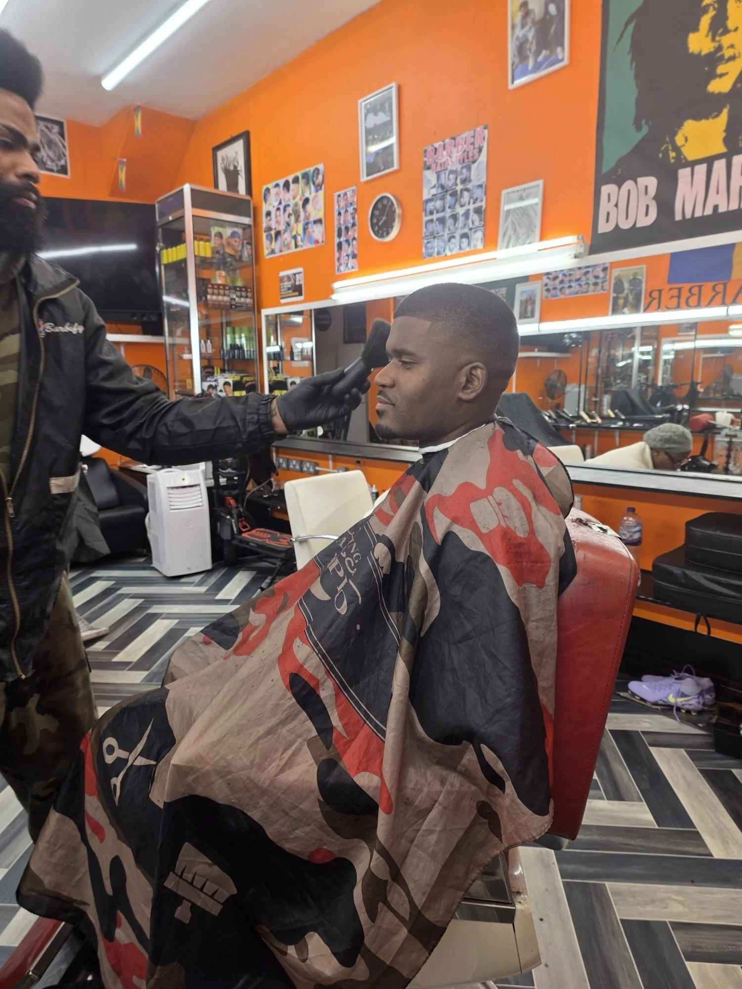 Man seated in a barber chair getting a fresh haircut, with a barber standing nearby. The barber is applying product on the man's face, possibly aftershave. The barbershop has orange walls decorated with framed pictures, posters, and a large Bob Marle