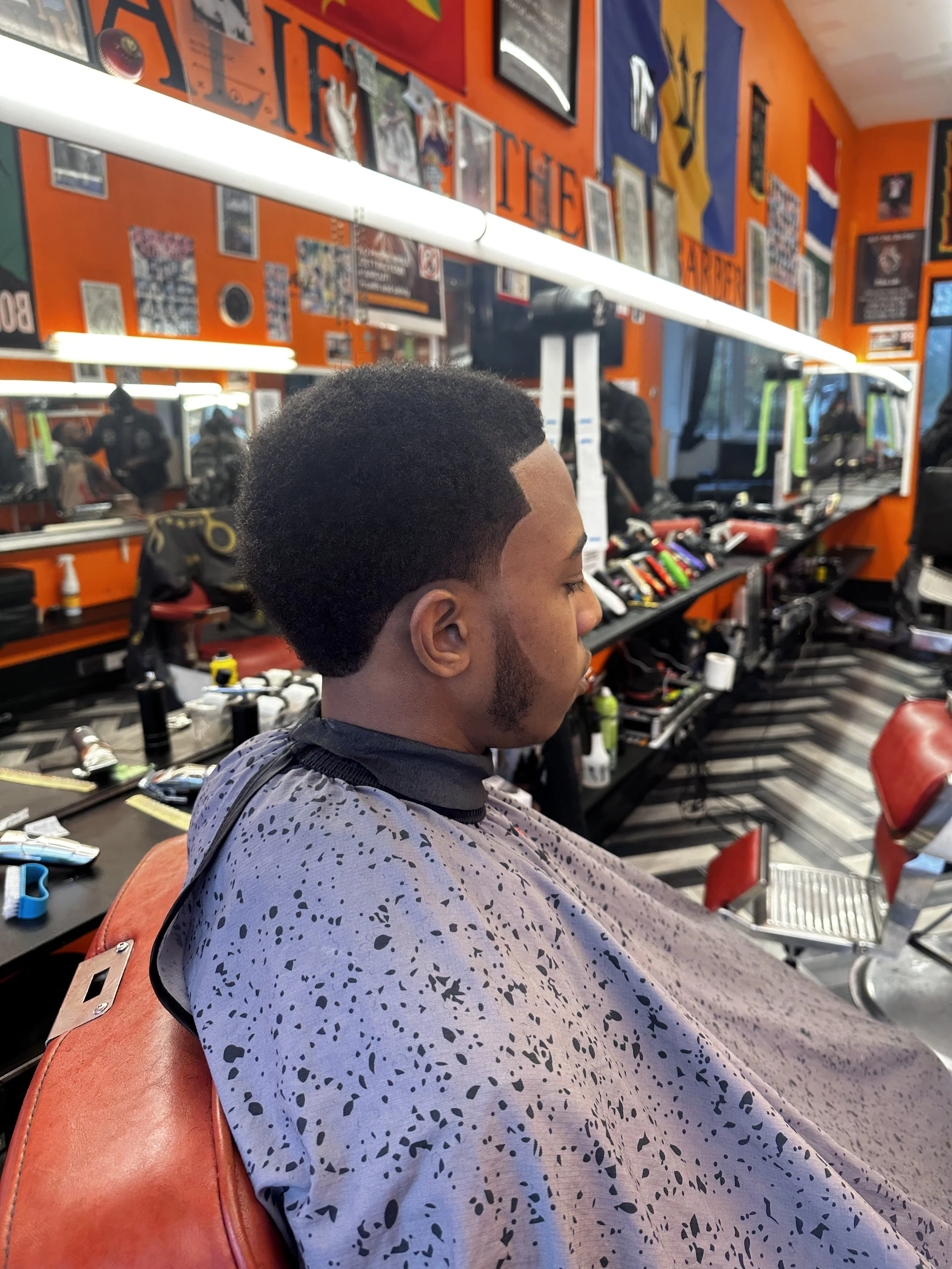 Young man sitting in a barber shop chair, getting a haircut, with mirrors and hair grooming tools visible in the background.