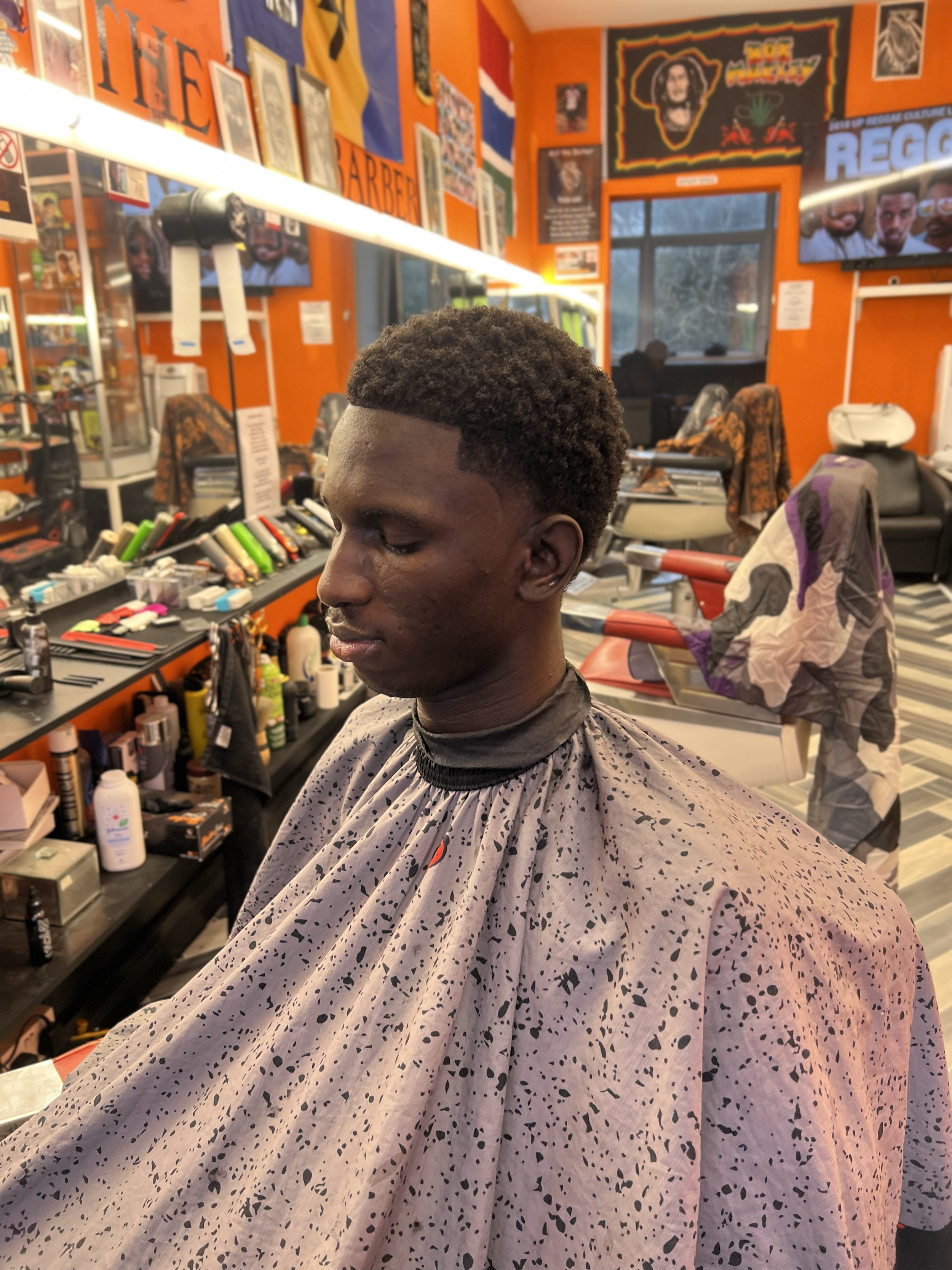 Young man getting a haircut in a barber shop with bright orange walls, posters, framed photos, and various barber tools visible.
