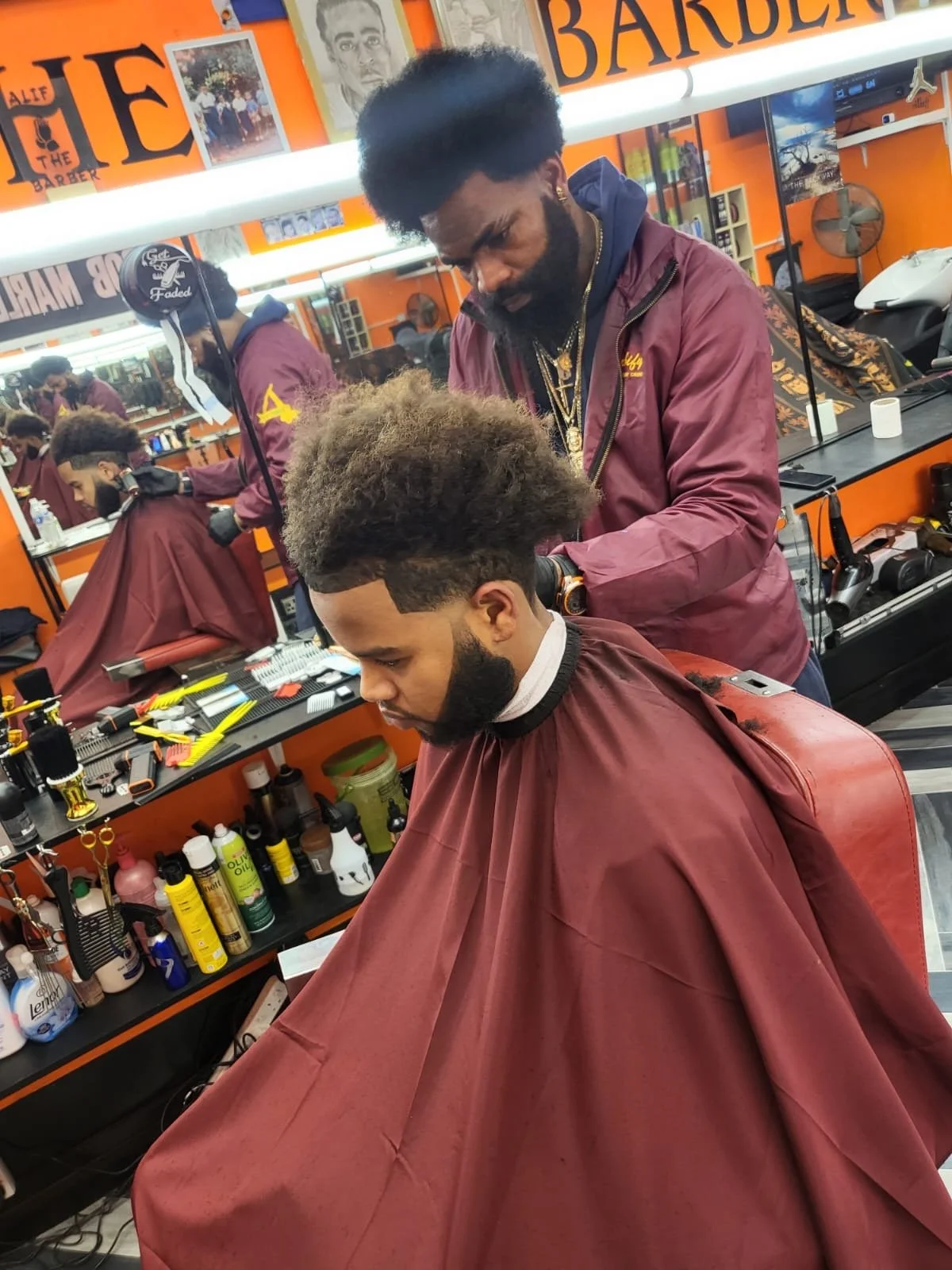 Barber giving a haircut in a barbershop with orange walls and signage. The barber is wearing a maroon jacket, and the customer has a fresh haircut with a beard and a large afro.