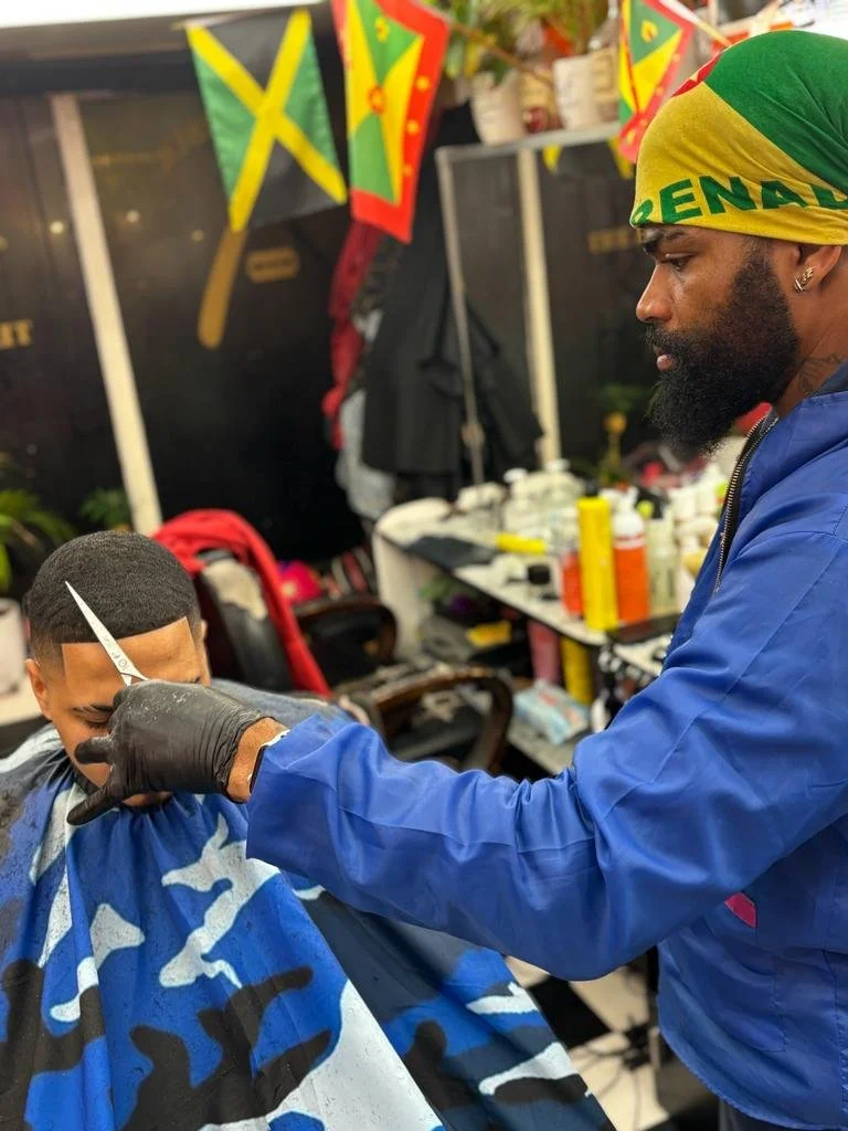 A man getting a haircut from a barber inside a barbershop adorned with Jamaican flags, with barber tools and products on the counter behind them.