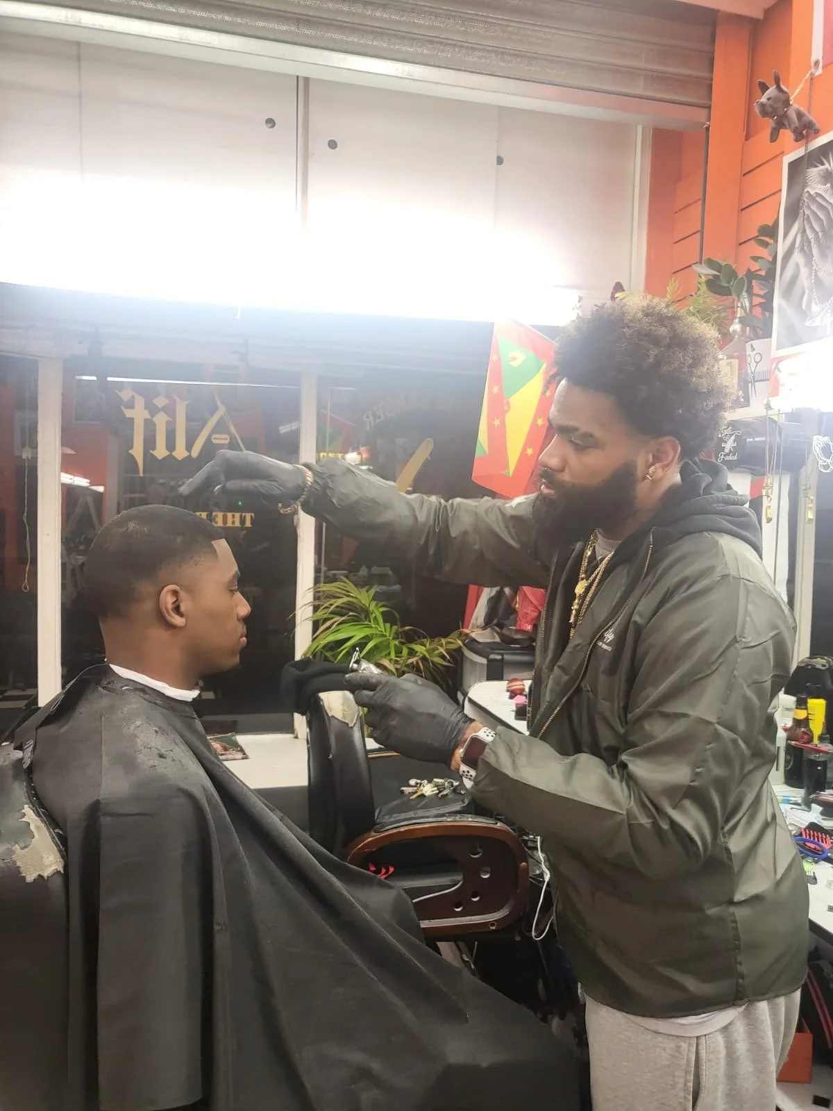 Barber giving a haircut to a young man in a barbershop with red walls and various decorations, including a dartboard and flags, and a plant on the counter.