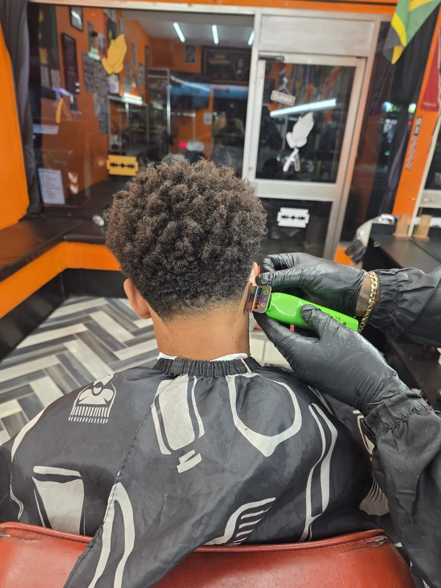 Person getting a haircut with a straight razor in a barbershop. The barber is wearing black gloves, and the person has short curly hair with a fade.